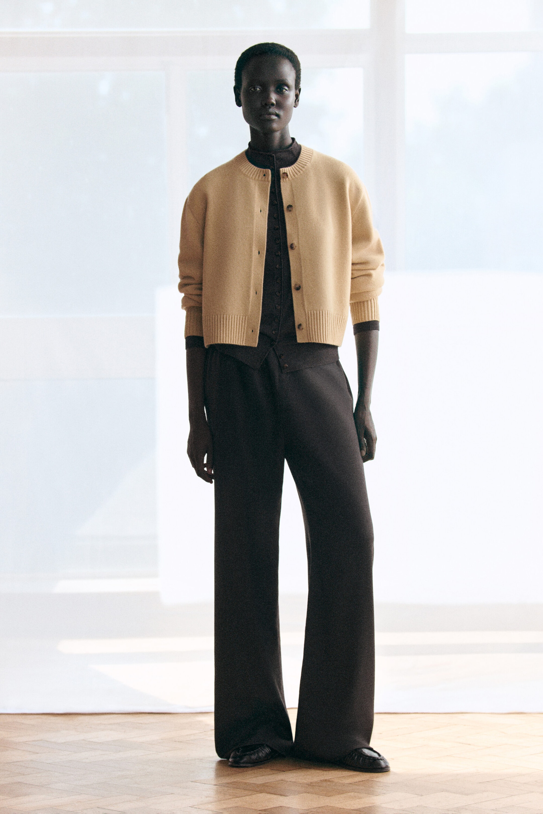 A cropped beige merino wool cardigan featuring a crew neck and ribbed trim is layered over a dark brown merino wool funnel-neck cardigan, both shown from the front to highlight their soft textures and relaxed fits. The dark brown funnel-neck cardigan is buttoned up and paired with matching wide-leg trousers, creating a coordinated, draped silhouette. A cropped beige merino wool cardigan featuring a crew neck and ribbed trim is layered over a dark brown merino wool funnel-neck cardigan, both shown from the front to highlight their soft textures and relaxed fits. The dark brown funnel-neck cardigan is buttoned up and paired with matching wide-leg trousers, creating a coordinated, draped silhouette.