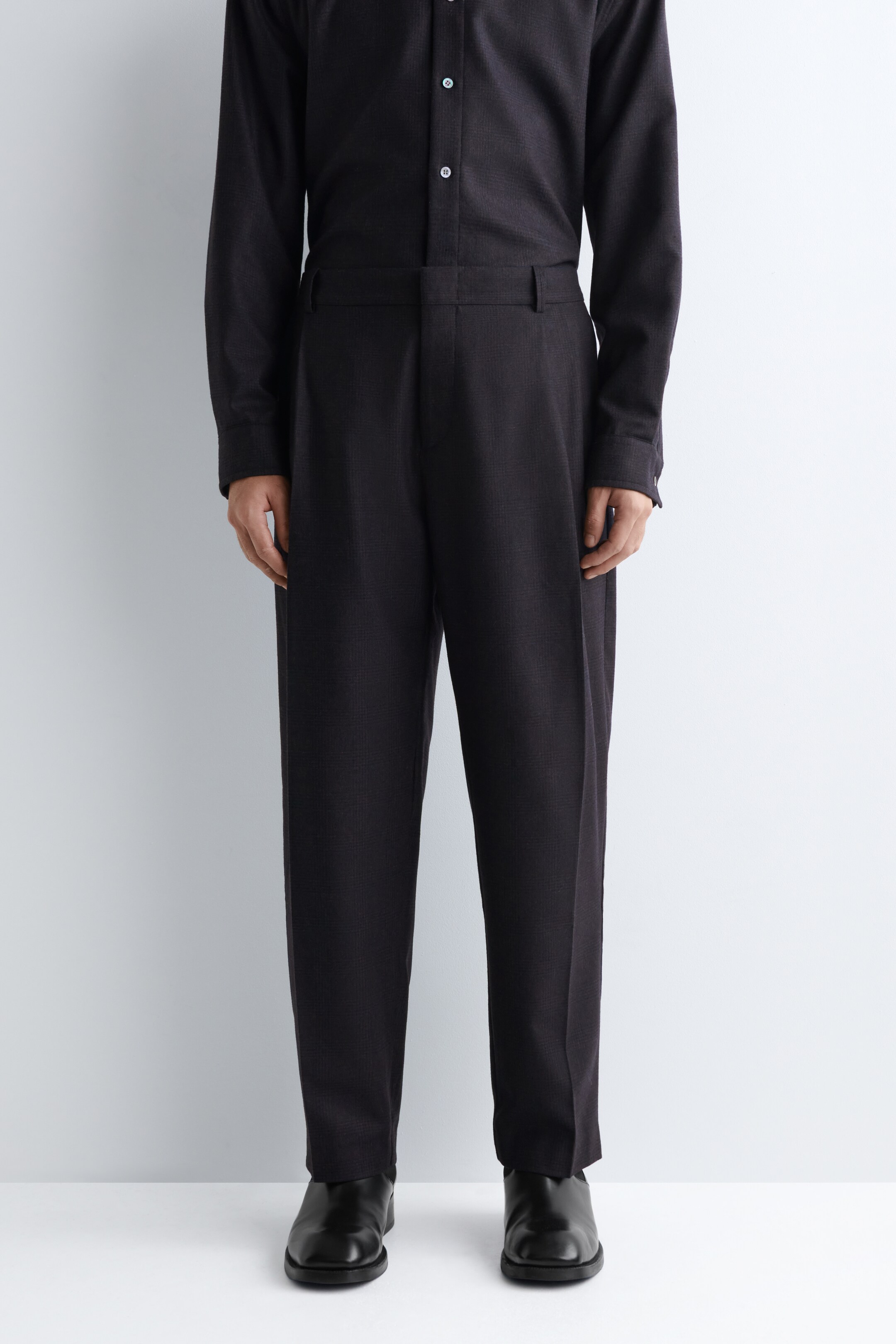 Agrandir l'image: Un ensemble sombre est porté, comprenant un pantalon large à taille haute avec des plis à l'avant, et une chemise à manches longues avec des boutons clairs. Les deux vêtements présentent un motif subtil et finement texturé. La tenue est orientée directement vers l'avant, complétée par des chaussures habillées noires.