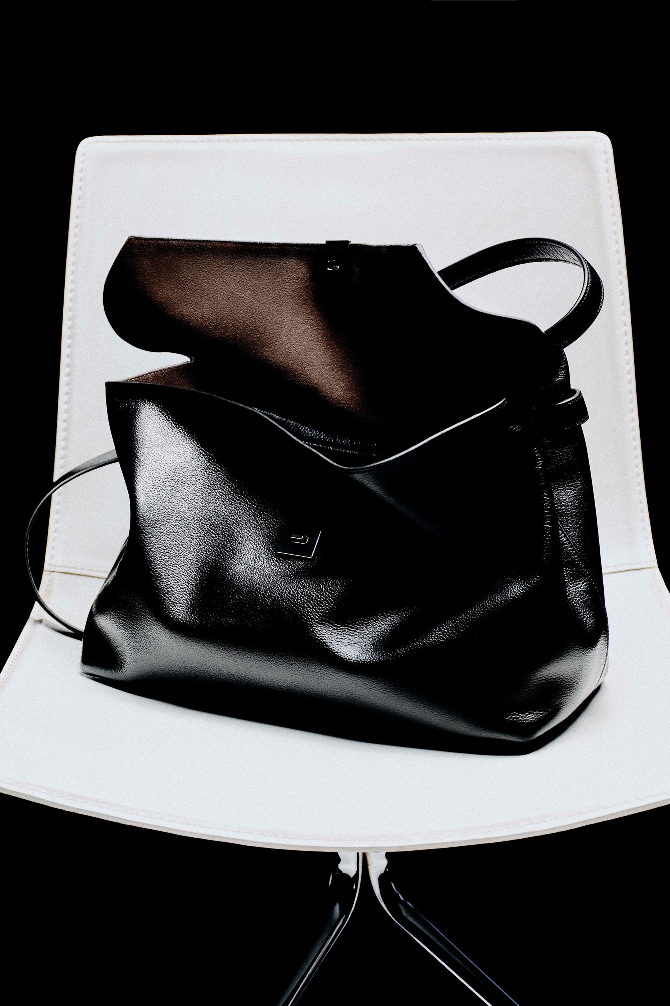 A front view of a black grained leather handbag is resting with its top flap open, revealing a dark interior lining and a soft, folded silhouette. The handbag is showcasing a small square metal clasp on the front, a top handle, and a thin shoulder strap draped to the side. A front view of a black grained leather handbag is resting with its top flap open, revealing a dark interior lining and a soft, folded silhouette. The handbag is showcasing a small square metal clasp on the front, a top handle, and a thin shoulder strap draped to the side.
