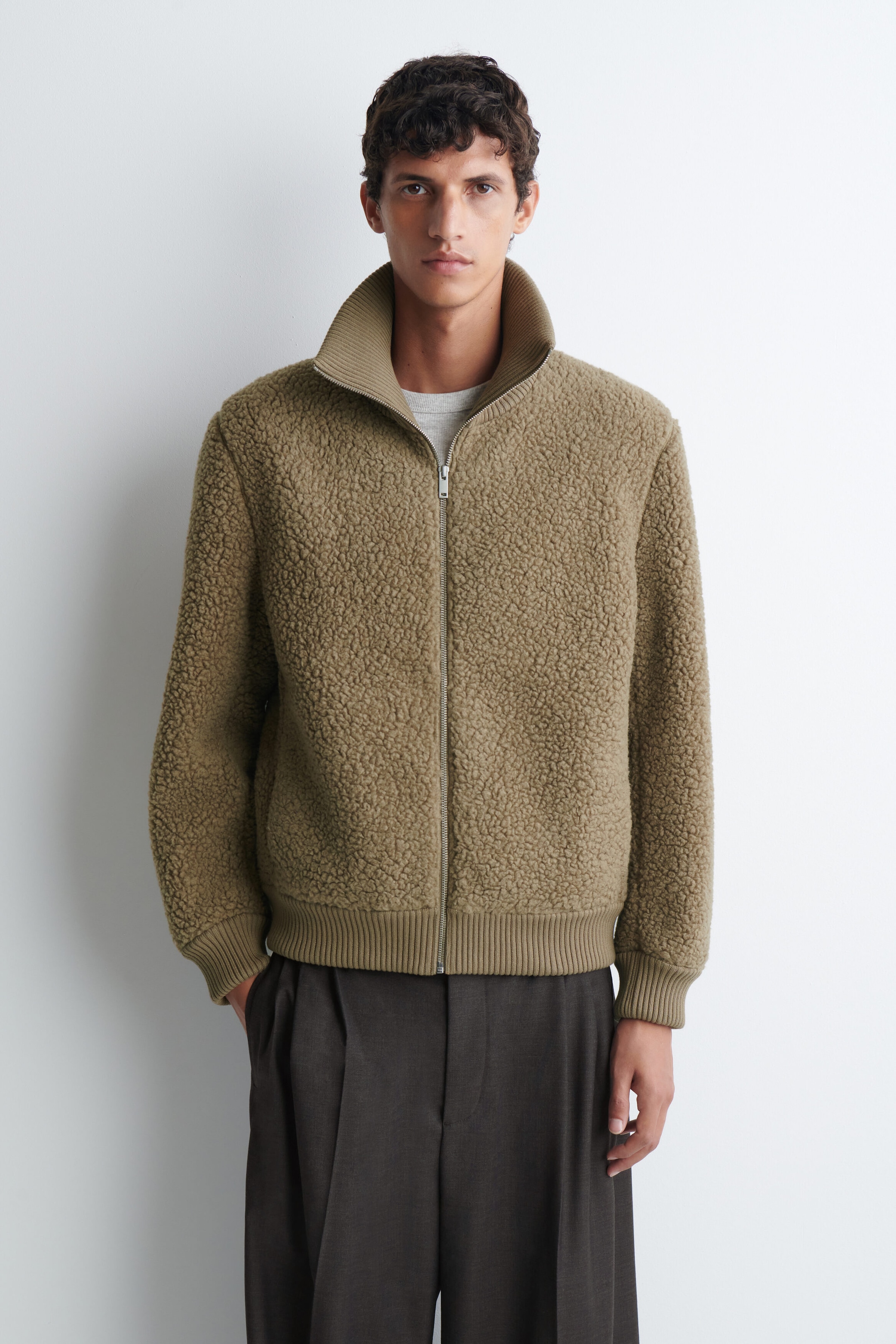 A beige-brown textured fleece jacket is being worn by the model, featuring a silver-tone front zipper, a high ribbed collar, and matching ribbed cuffs and hem. The jacket is layered over a grey crew-neck T-shirt and paired with dark brown pleated trousers. A beige-brown textured fleece jacket is being worn by the model, featuring a silver-tone front zipper, a high ribbed collar, and matching ribbed cuffs and hem. The jacket is layered over a grey crew-neck T-shirt and paired with dark brown pleated trousers.