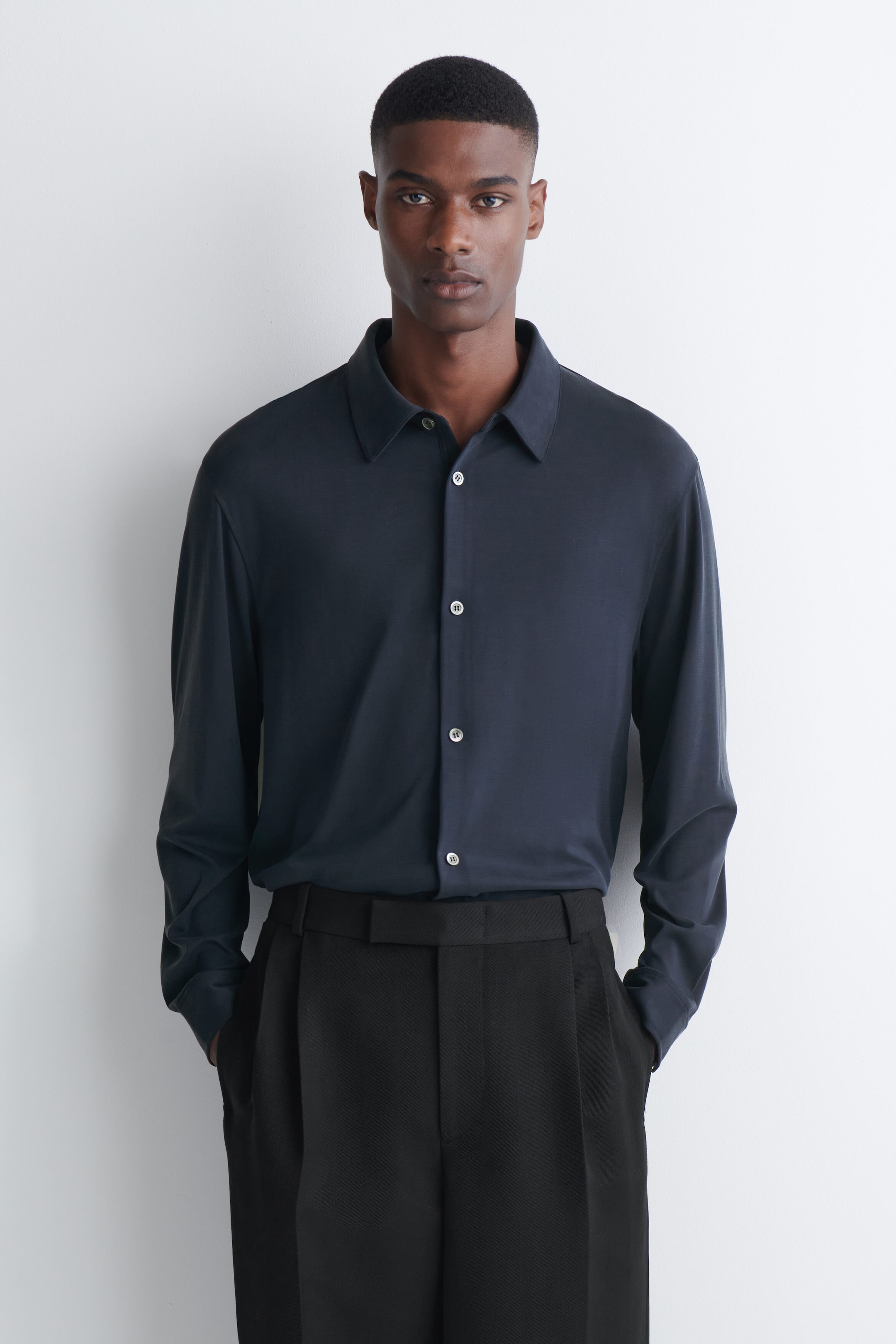 The model is wearing a fluid interlock jersey shirt in dark navy blue, tucked into black trousers. The long-sleeved shirt features a pointed collar and a front button closure with light-colored buttons. The model is wearing a fluid interlock jersey shirt in dark navy blue, tucked into black trousers. The long-sleeved shirt features a pointed collar and a front button closure with light-colored buttons.