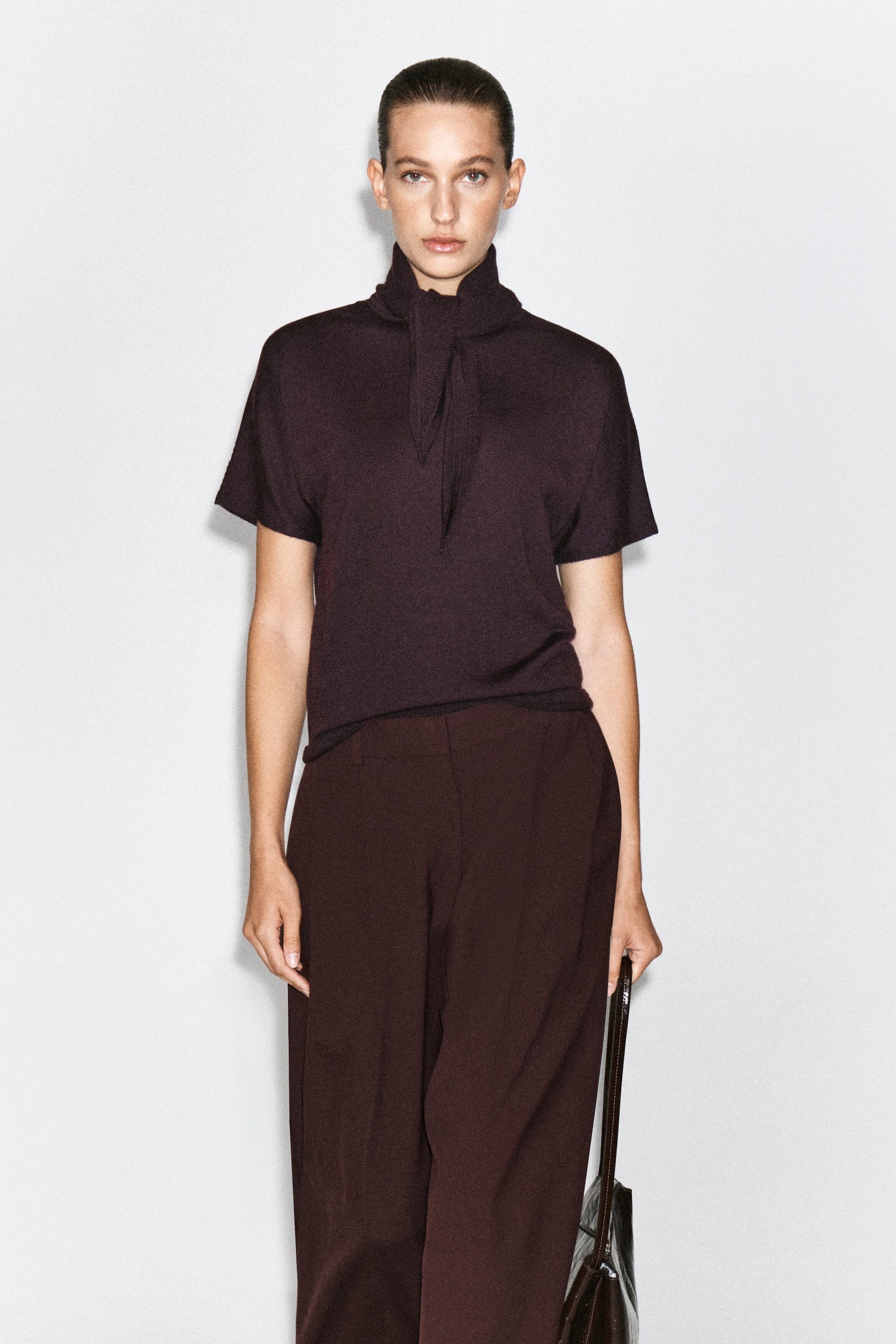 The scarf-detail merino wool blend T-shirt in dark plum is worn by the model and features short sleeves and a high collar with an integrated draped front tie. The T-shirt is paired with matching dark trousers, and a dark leather handbag is carried in one hand.