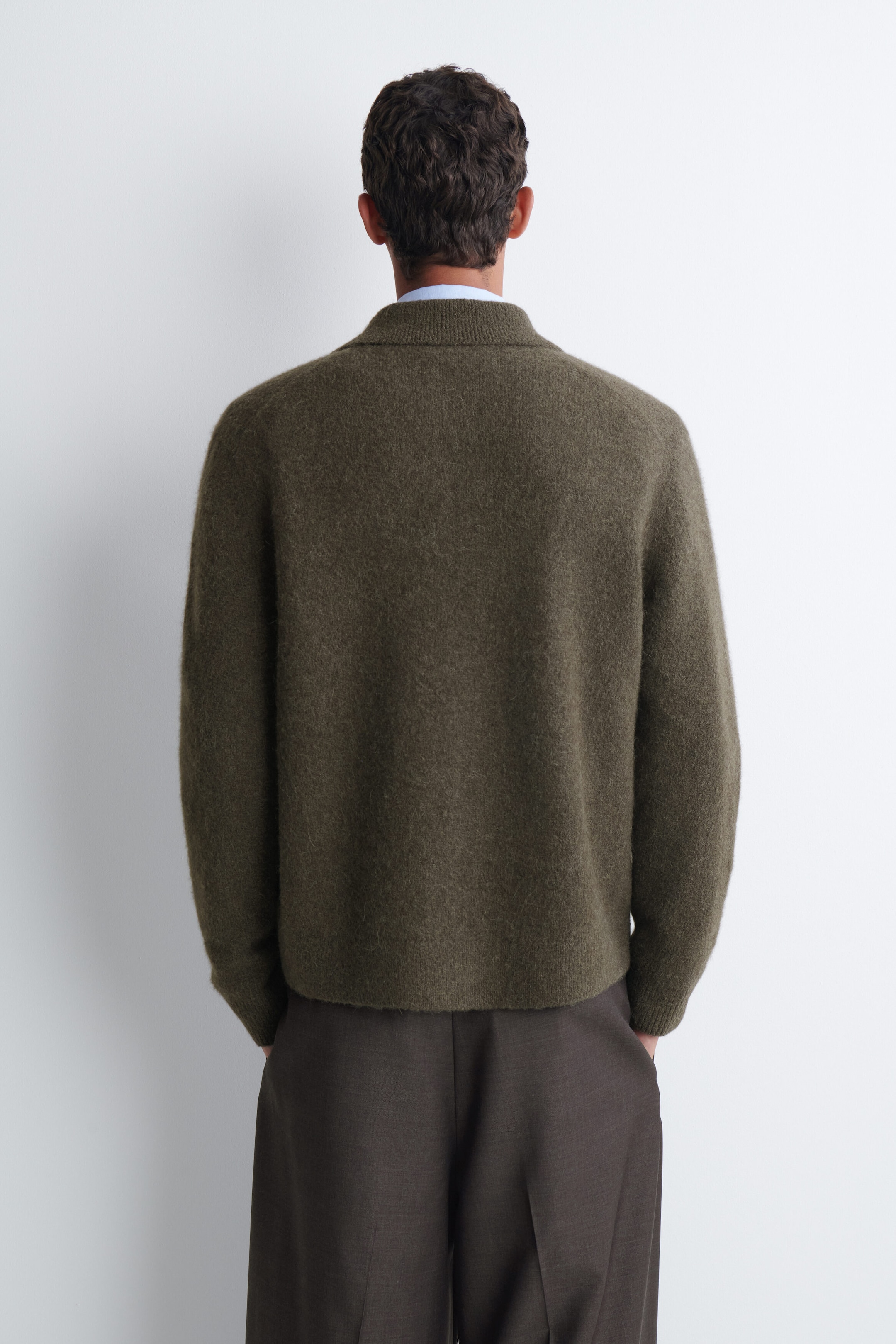 A textured collared cardigan in olive green is being worn by the model, shown from the back. The cardigan features a soft, fuzzy knit texture and is layered over a light blue collared shirt and paired with dark brown trousers. A textured collared cardigan in olive green is being worn by the model, shown from the back. The cardigan features a soft, fuzzy knit texture and is layered over a light blue collared shirt and paired with dark brown trousers.