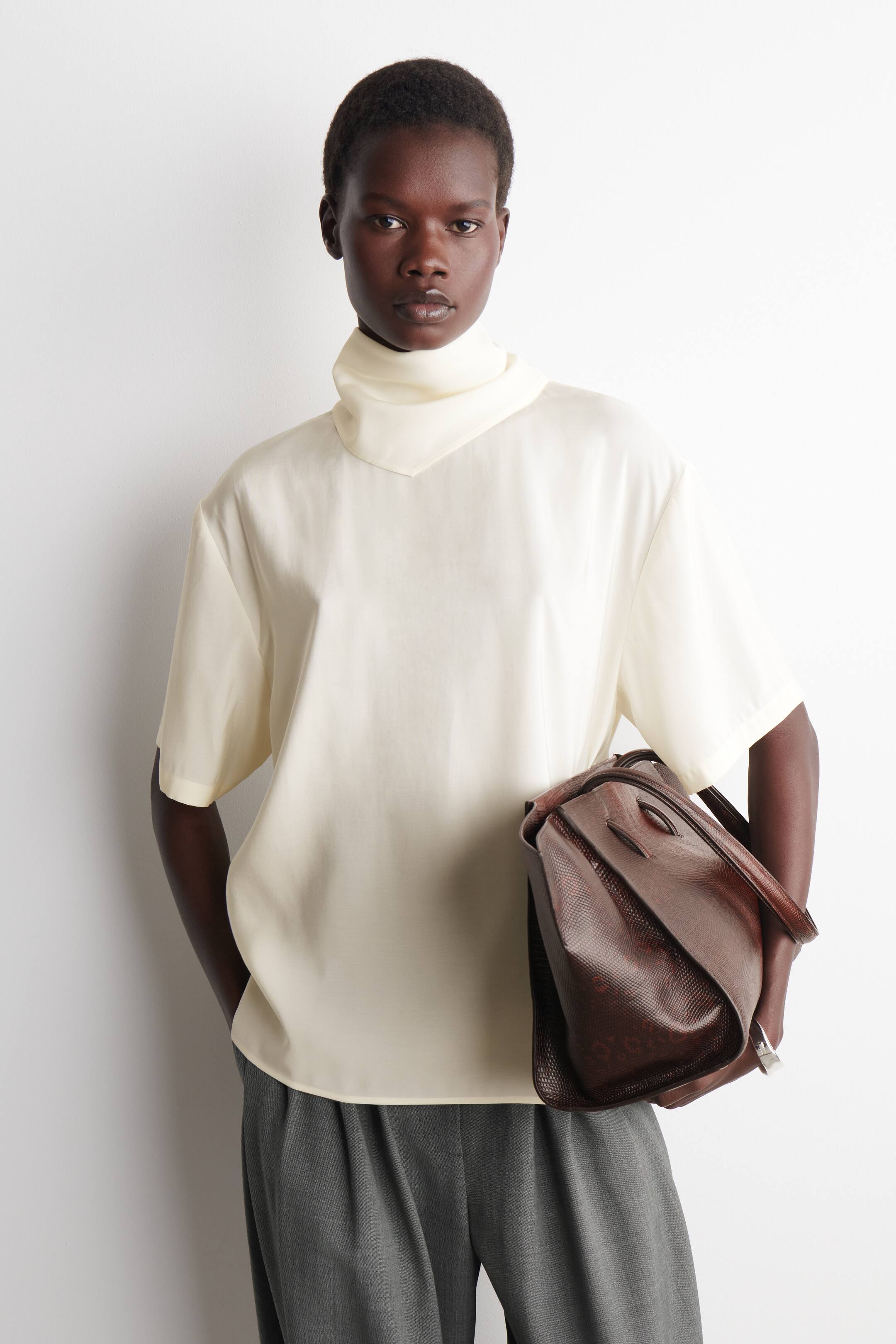 Agrandir l'image: Un sac à main marron foncé est présenté en vue trois-quarts, mettant en valeur sa forme rectangulaire structurée. Il est doté de deux poignées supérieures, d'un petit rabat de fermeture et d'une texture à effet lézard distincte.