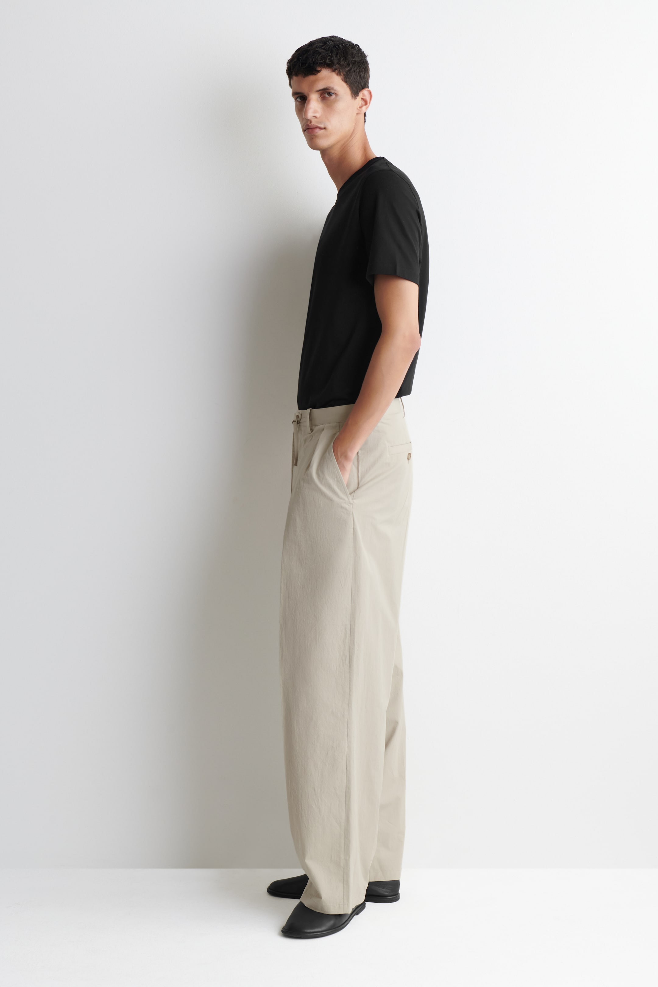 Grotere afbeelding bekijken: Een zwart T-shirt met ronde hals wordt gedragen, ingestopt in licht beige wijde broek. De broek wordt getoond vanuit een zij-voorhoek, met voorplooien en zijzakken, waarbij één hand in een zak rust, en reikt tot op de vloer over zwarte schoenen.