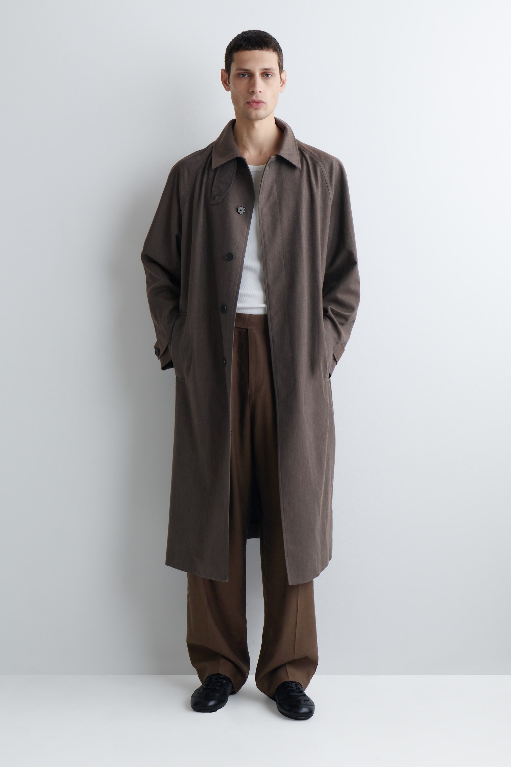 Zobraziť väčší obrázok: Tmavohnedý jednoradový dlhý kabát je otočený dopredu, s jemnou vertikálnou textúrou, rozšíreným golierom, raglánovými rukávmi a dĺžkou po polovicu lýtok, vo voľnom strihu. Je nosený otvorený cez biele tričko, odhaľujúc tri tmavé gombíky a bočné šikmé vrecká.