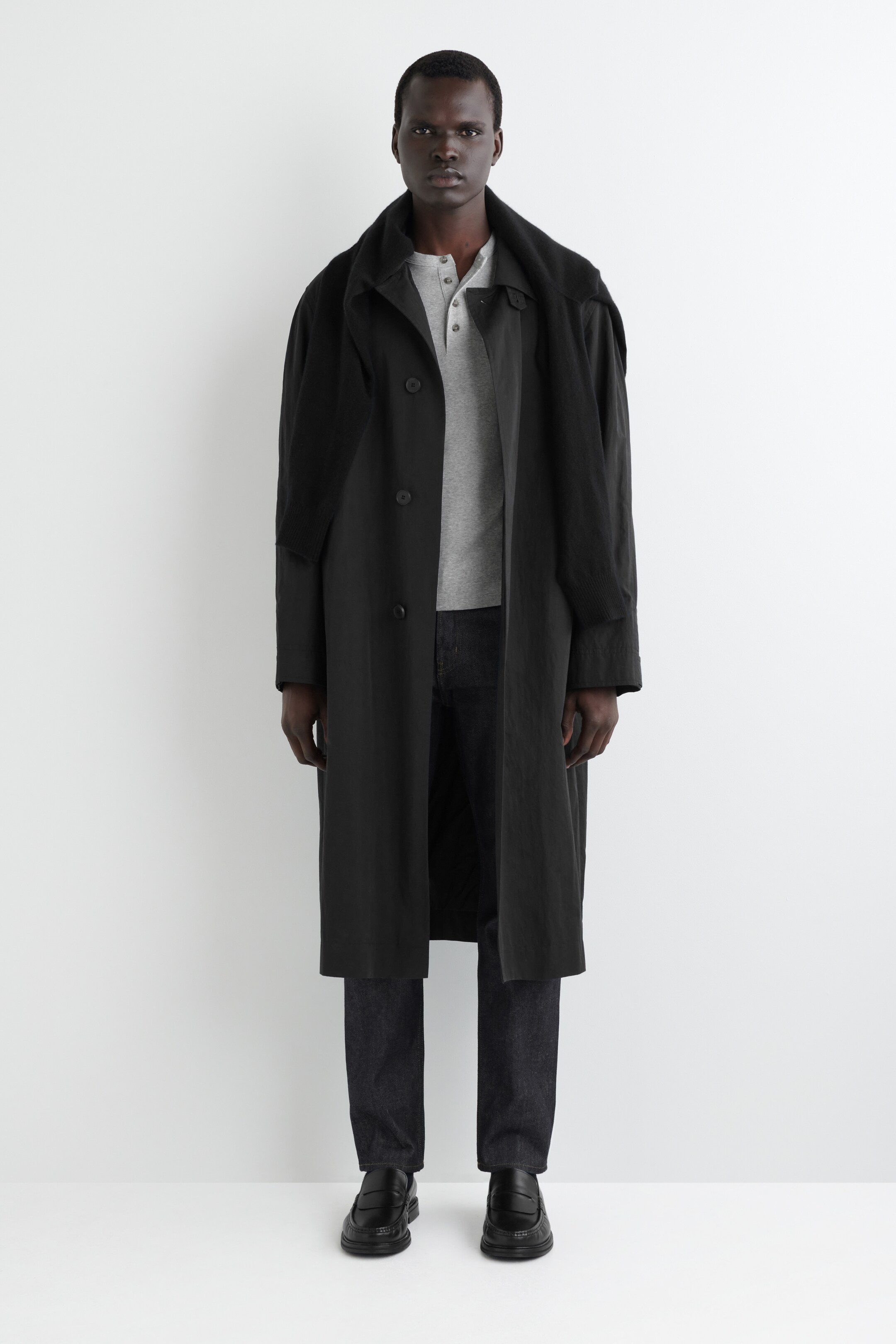 Agrandir l'image: Un manteau noir à simple boutonnage est présenté de face. Le manteau présente une coupe décontractée, un col classique et descend sous les genoux. Il est porté ouvert sur un t-shirt Henley gris, avec un vêtement texturé et sombre drapé sur ses épaules.