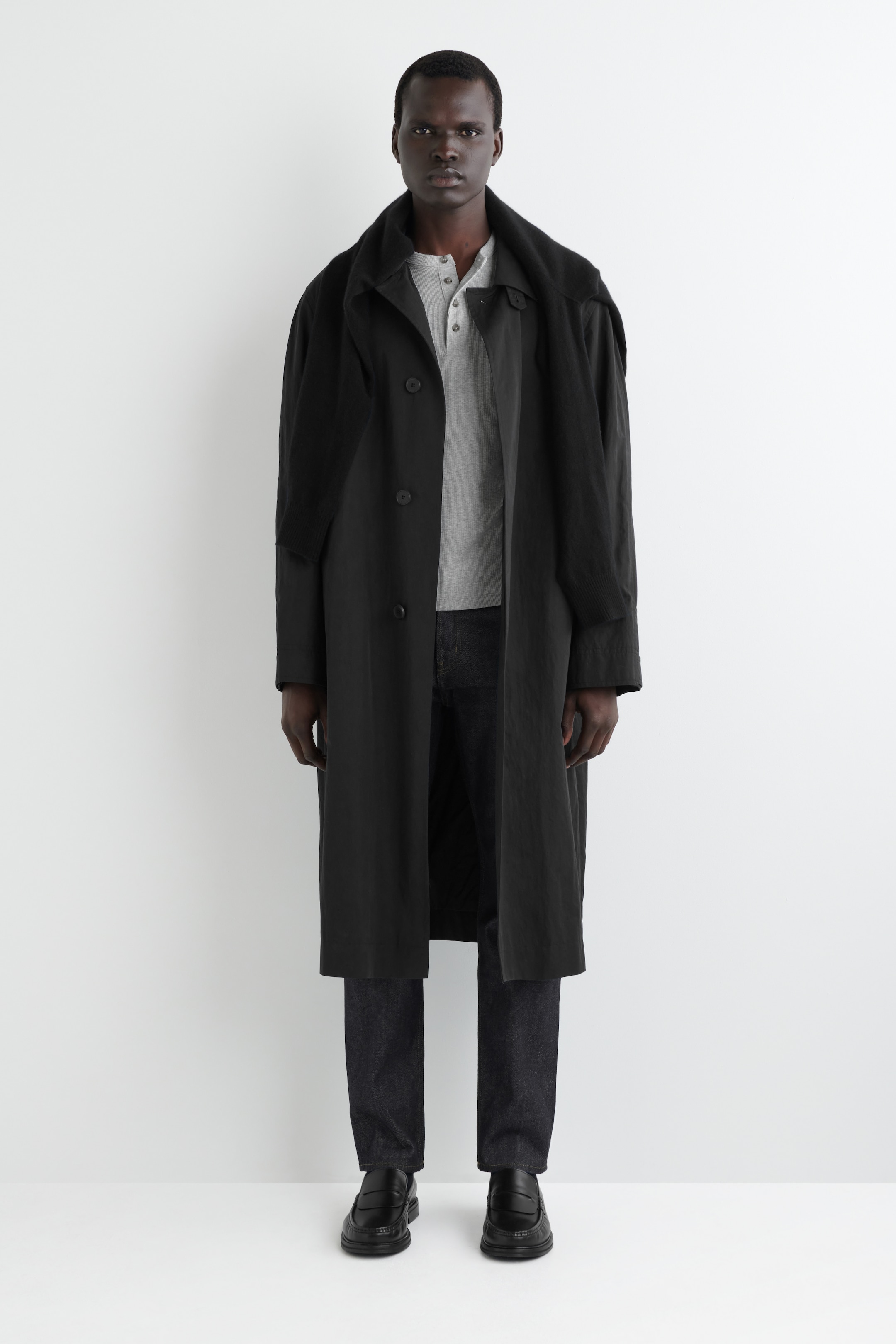 View larger image: A black, single-breasted coat is being presented from a full frontal angle. The coat features a relaxed fit, a classic collar, and is reaching below the knees. It is being styled open over a grey Henley shirt, with a dark, textured garment draped across its shoulders.