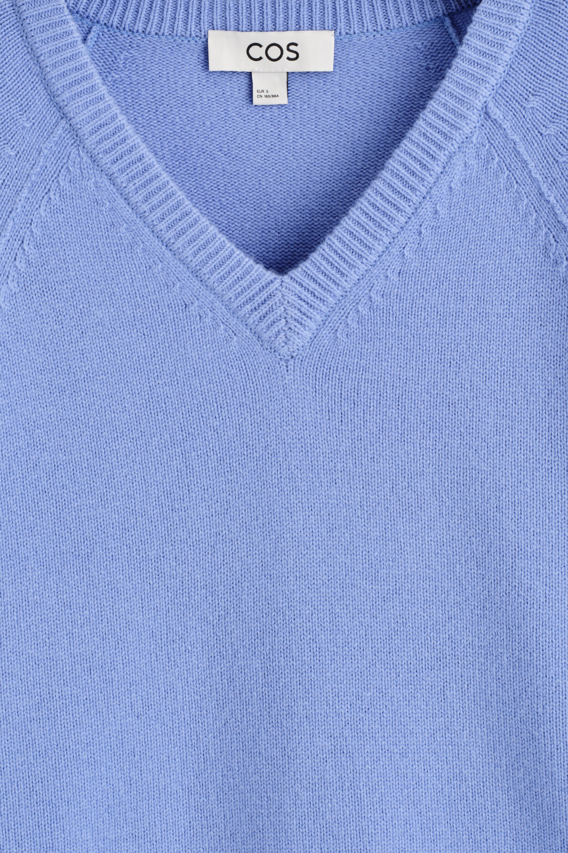 Ingrandisci l'immagine: Un maglione a maglia azzurro chiaro con scollo a V è mostrato dal DAVANTI, con uno scollo a V a coste e cuciture delle maniche raglan visibili. Un'etichetta bianca del marchio COS è visibile sul retro del collo.