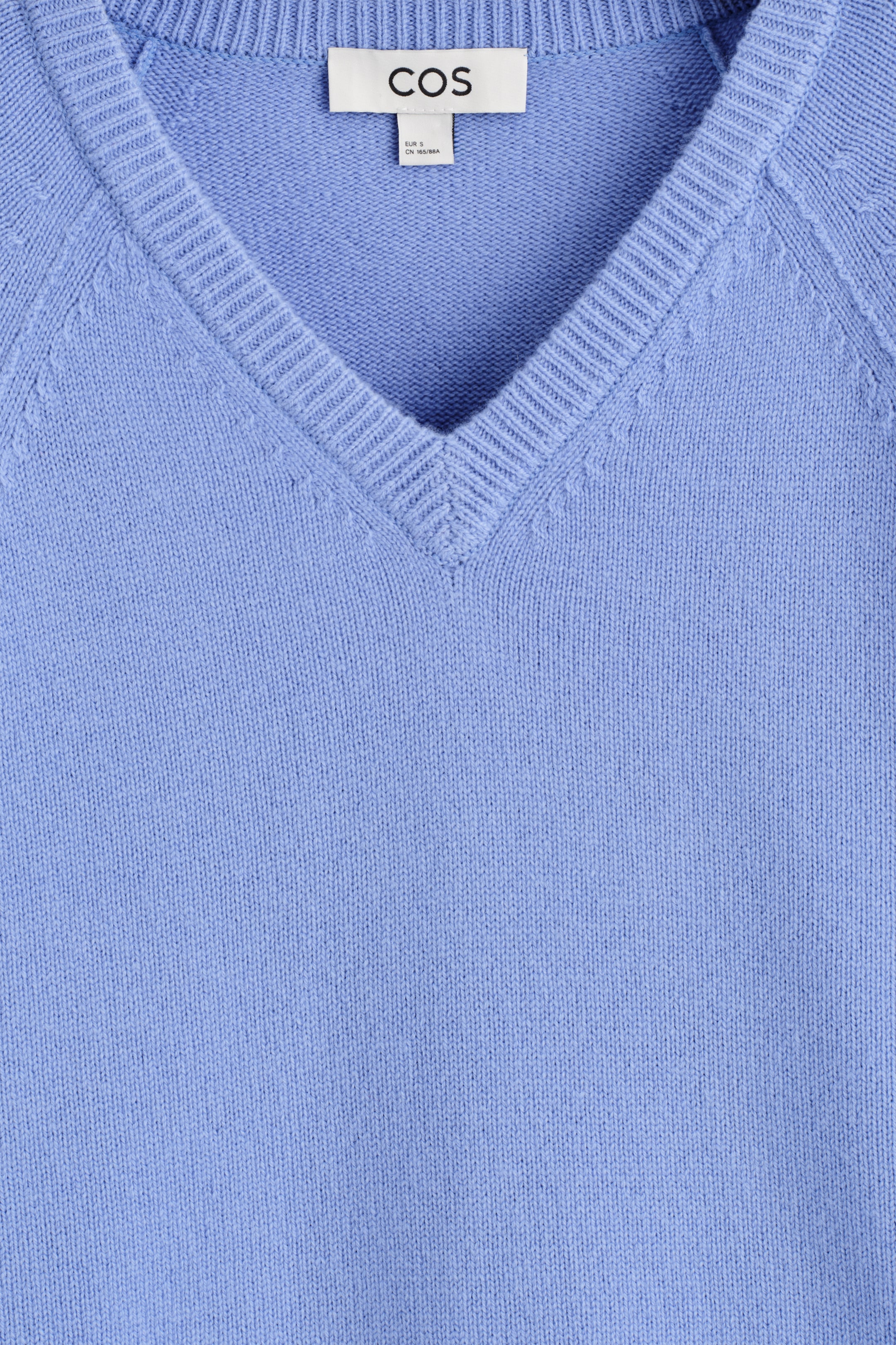 Visualizza immagine più grande: Un maglione a maglia azzurro chiaro con scollo a V è mostrato dal DAVANTI, con uno scollo a V a coste e cuciture delle maniche raglan visibili. Un'etichetta bianca del marchio COS è visibile sul retro del collo.