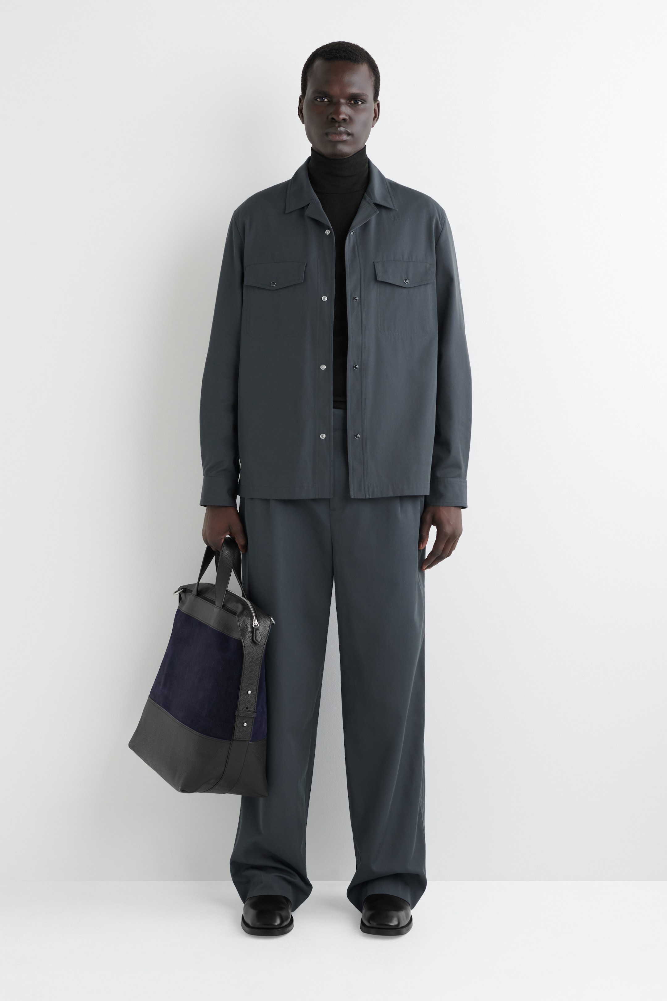 View larger image: A dark grey shirt jacket is being presented full-front, featuring a spread collar, two buttoned chest pockets, and a front button closure, and is layered over a black turtleneck. Matching dark grey wide-leg trousers are completing the ensemble, with black dress shoes and a two-toned dark grey and navy handbag being held.