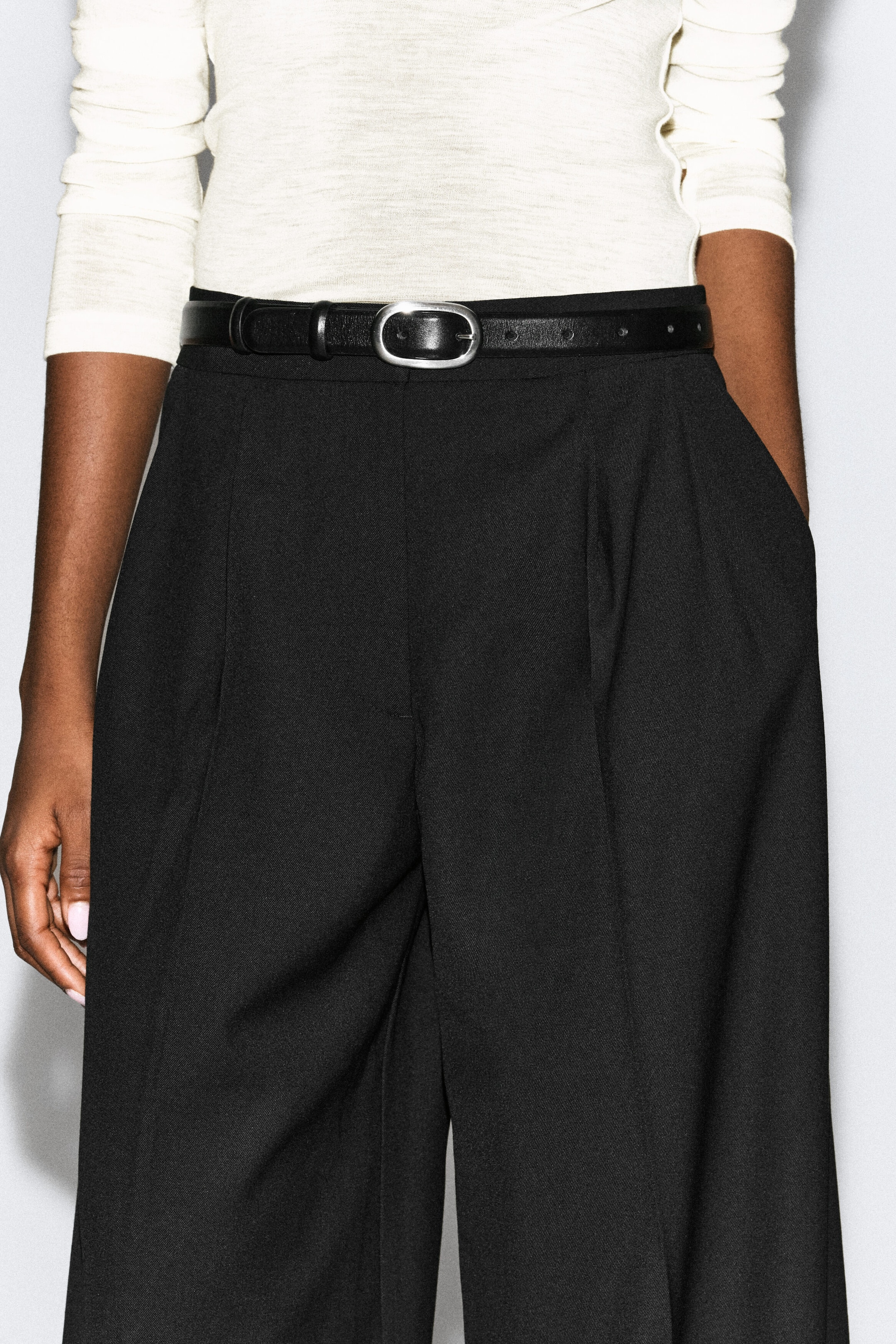 The model is wearing pleated wide-leg pants in black, cinched at the waist with a thin black leather belt featuring a silver-tone oval buckle. The high-waisted pants are styled with a tucked-in, cream-colored, long-sleeved top. The model is wearing pleated wide-leg pants in black, cinched at the waist with a thin black leather belt featuring a silver-tone oval buckle. The high-waisted pants are styled with a tucked-in, cream-colored, long-sleeved top.