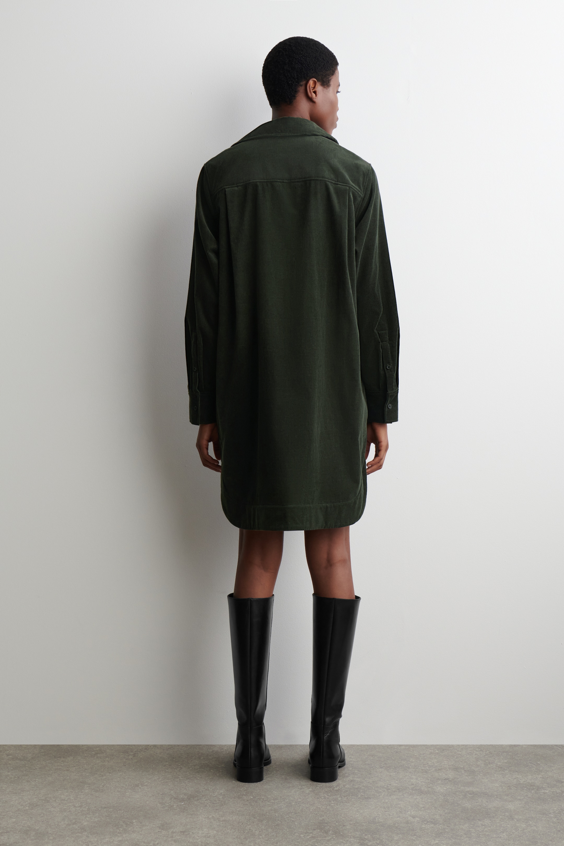 Visualizza immagine più grande: Un abito camicia di velluto a coste verde scuro viene presentato dal retro, con una carré sulla parte superiore della schiena, maniche lunghe e un orlo arrotondato che cade sopra il ginocchio. Viene abbinato a stivali neri alti fino al ginocchio.
