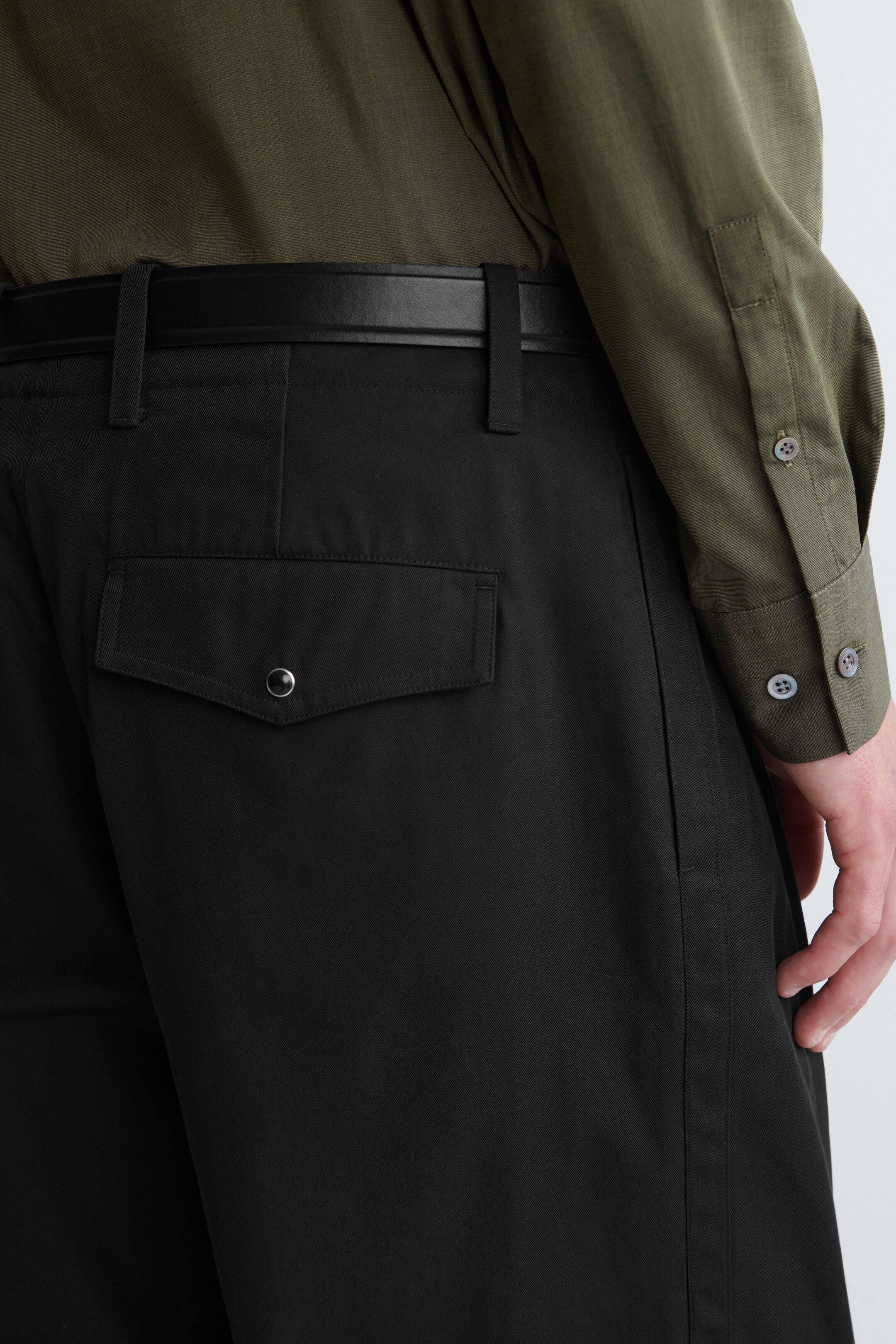 Visualizza immagine più grande: I pantaloni neri presentano una cintura nera infilata nei passanti e una camicia verde oliva infilata nella cintura, vista da un angolo posteriore. Una tasca posteriore con patta appuntita e chiusura a scatto color argento è visibile sul lato destro.