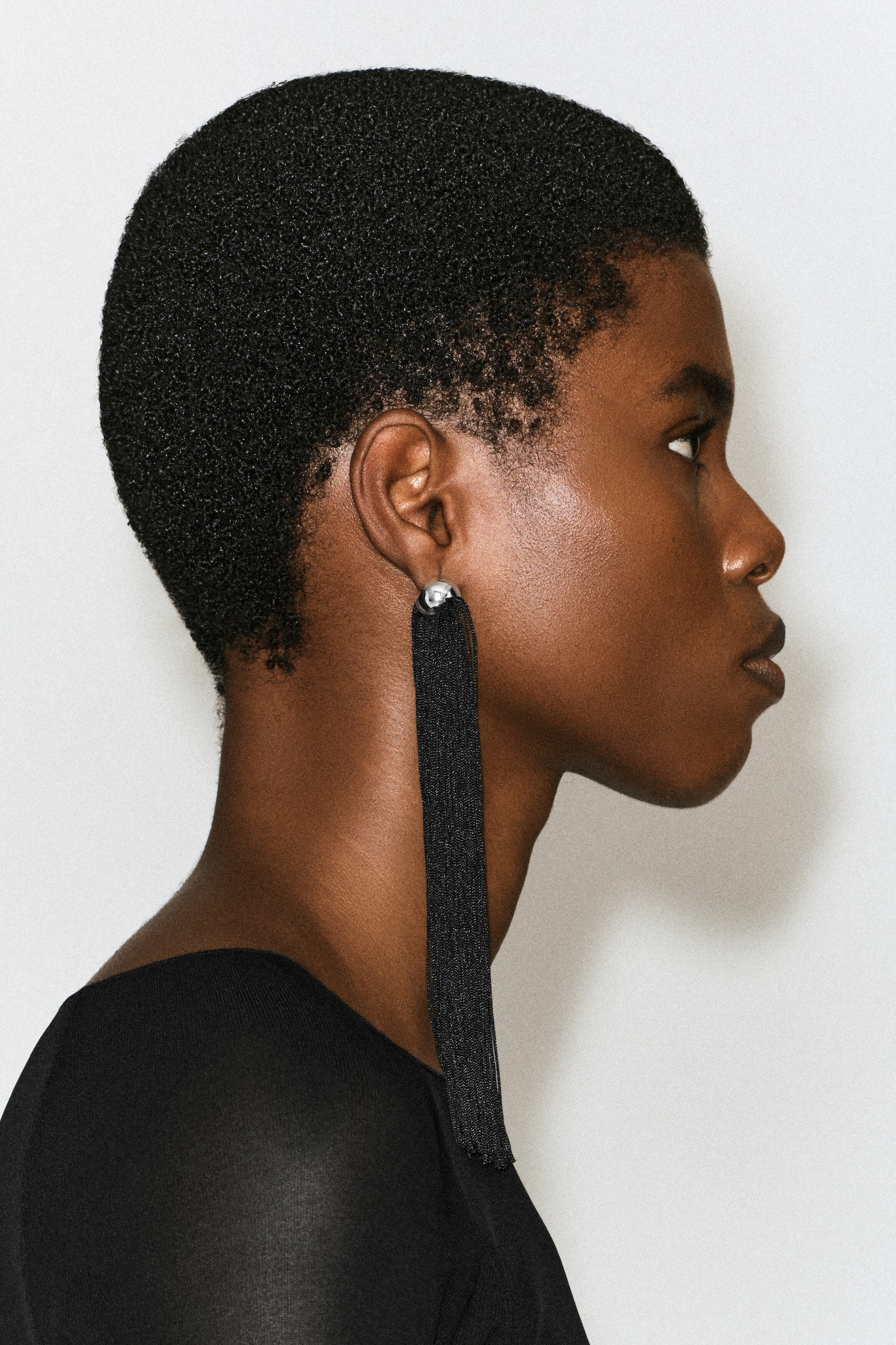 A fringed earring featuring a silver-tone spherical stud and a long, dense tassel of fine black threads is worn on the model's earlobe. The long black fringe hangs below the neck, draped over a black garment.