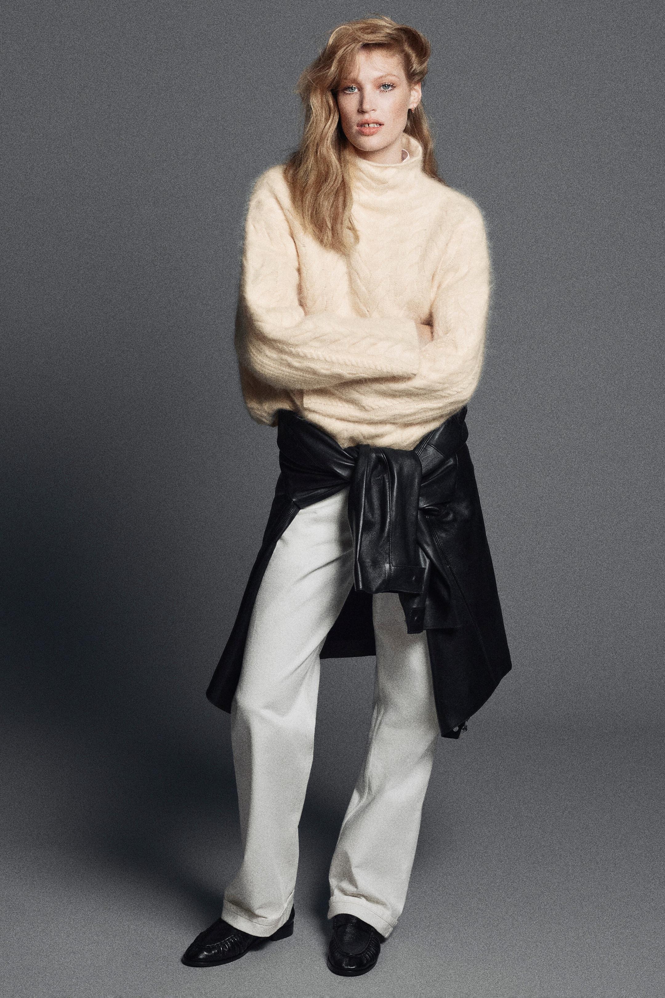 In a front-facing view, a model is wearing a cream-colored cable-knit brushed-cashmere funnel-neck sweater featuring a textured, relaxed fit, paired with off-white curved-leg turn-up jeans. A black leather jacket is wrapped around the waist over the jeans, and black loafers complete the styled look. In a front-facing view, a model is wearing a cream-colored cable-knit brushed-cashmere funnel-neck sweater featuring a textured, relaxed fit, paired with off-white curved-leg turn-up jeans. A black leather jacket is wrapped around the waist over the jeans, and black loafers complete the styled look.