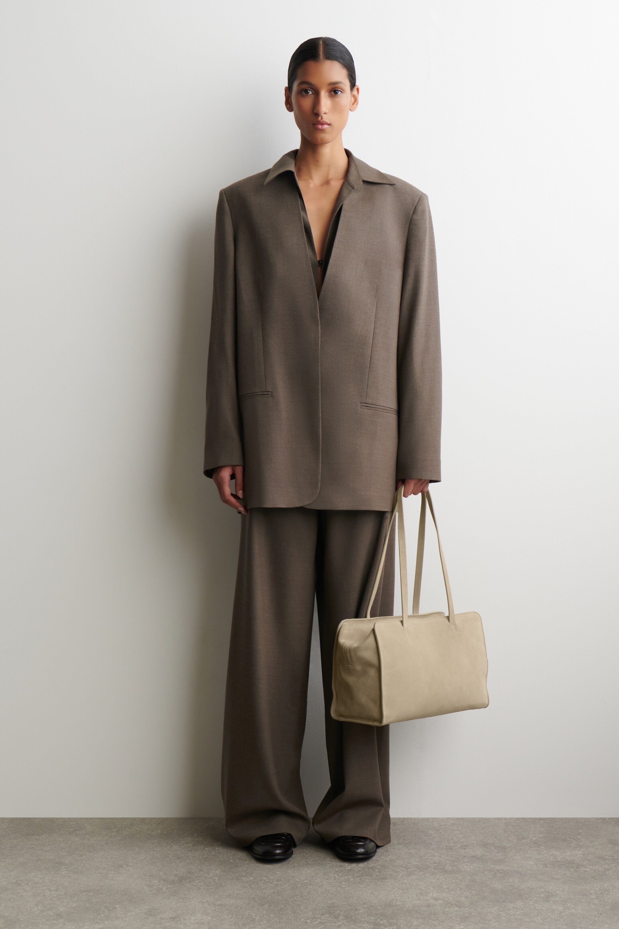 Zobrazit větší obrázek: Tmavě béžový oversized oblek, který obsahuje jednořadé sako s klasickým límcem, širokými rameny a klopovými kapsami, je předváděn s odpovídajícími kalhotami s širokými nohavicemi. Světle béžová obdélníková kabelka přes rameno se dvěma plochými popruhy je držena a jsou nošeny černé ploché boty.