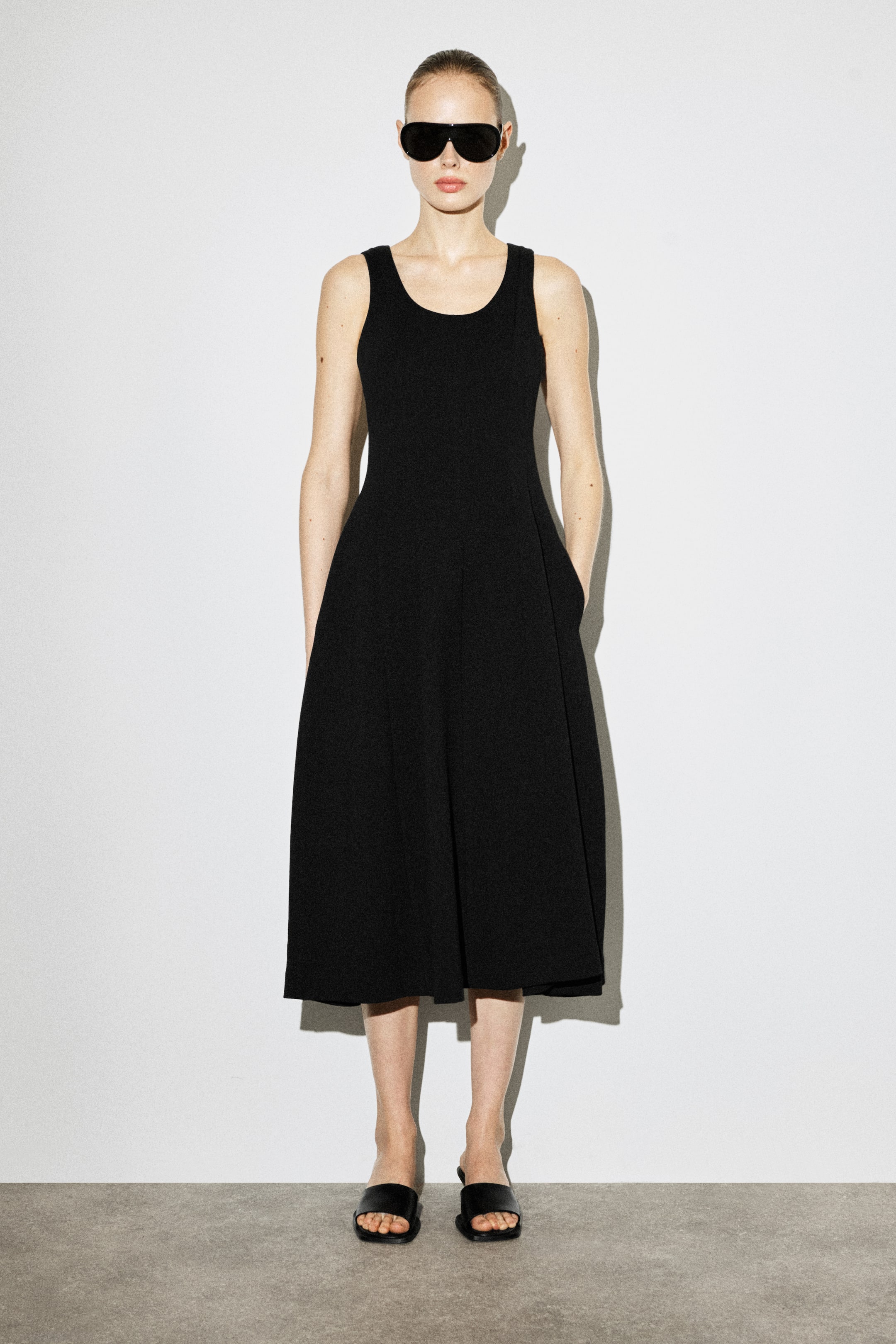 Zobraziť väčší obrázok: Čierne midi šaty bez rukávov majú okrúhly výstrih, priliehavý živôtik a sukňu v tvare A, ktorá siaha do midi dĺžky. Šaty majú bočné vrecká, do ktorých sú vložené ruky.