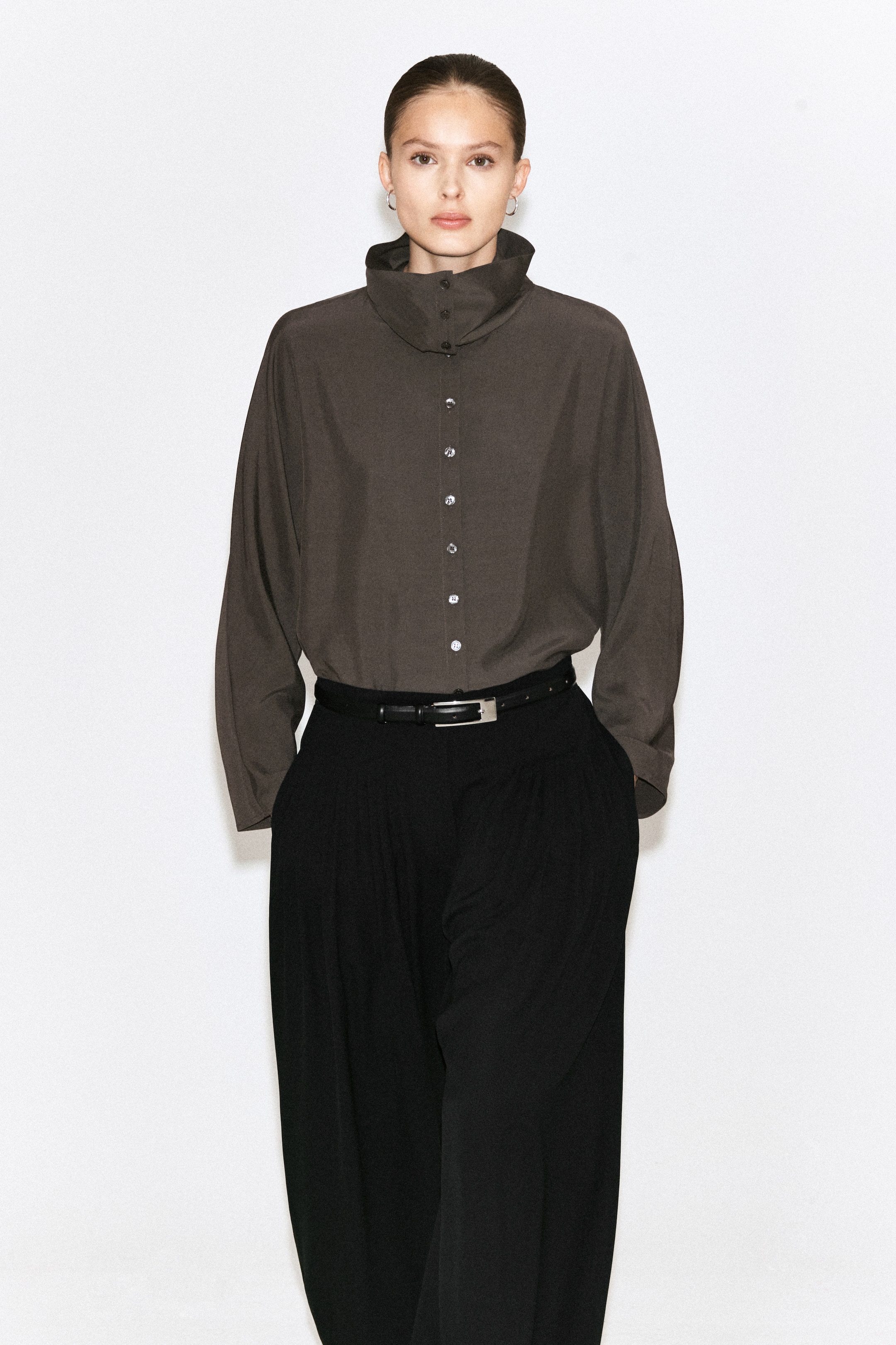 Ver imagen más grande: Una camisa de manga larga de color marrón oscuro, con un cuello alto abotonado y una tapeta completa de botones, se lleva metida dentro de unos pantalones negros de pierna ancha con pliegues. Los pantalones están ceñidos en la cintura por un cinturón negro con una hebilla plateada.