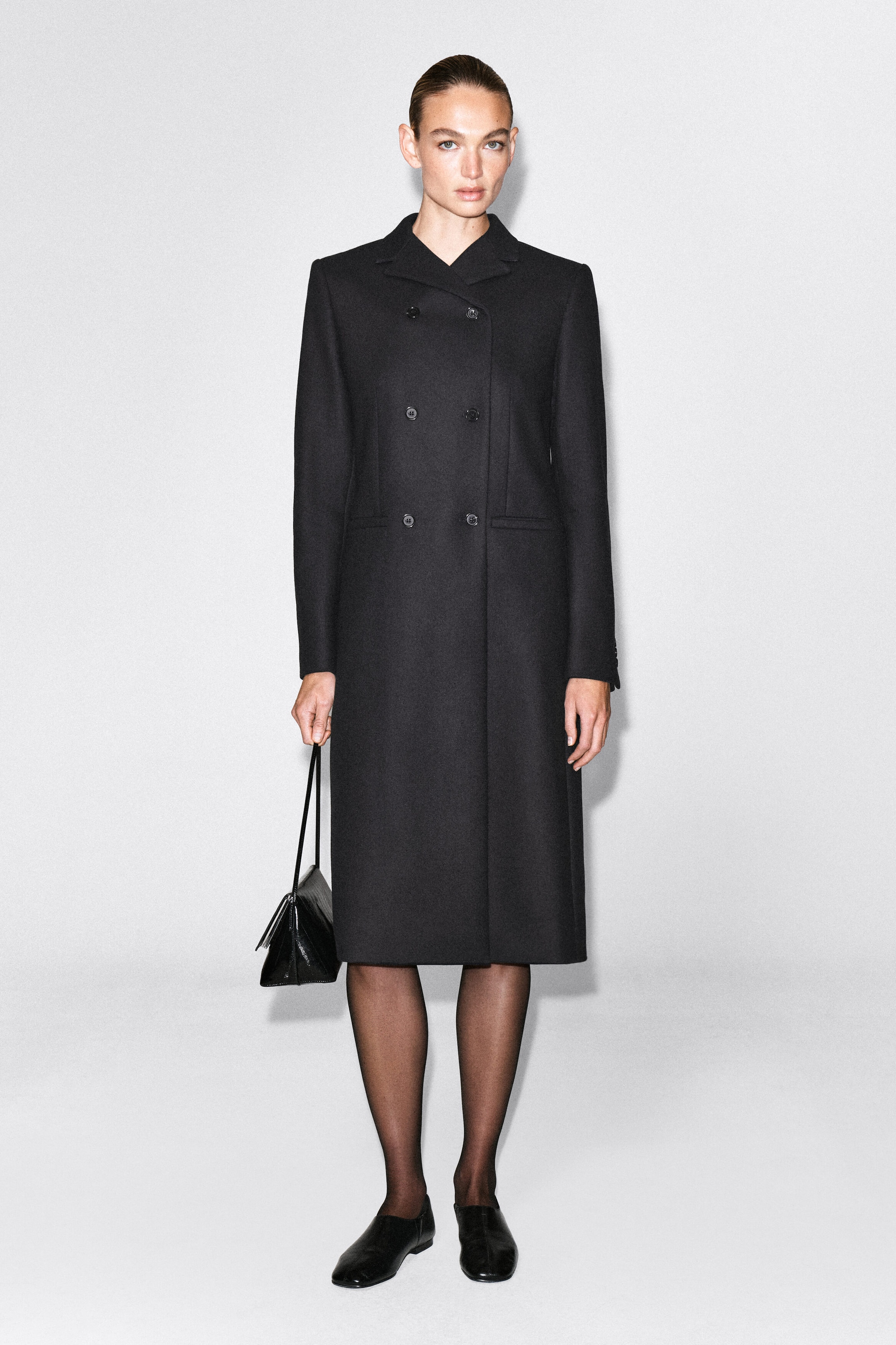 The model is wearing a longline, double-breasted black coat featuring a slim, structured silhouette, classic collar, and button closures. She styles the coat with sheer black tights, black flats, and a small black handbag. The model is wearing a longline, double-breasted black coat featuring a slim, structured silhouette, classic collar, and button closures. She styles the coat with sheer black tights, black flats, and a small black handbag.