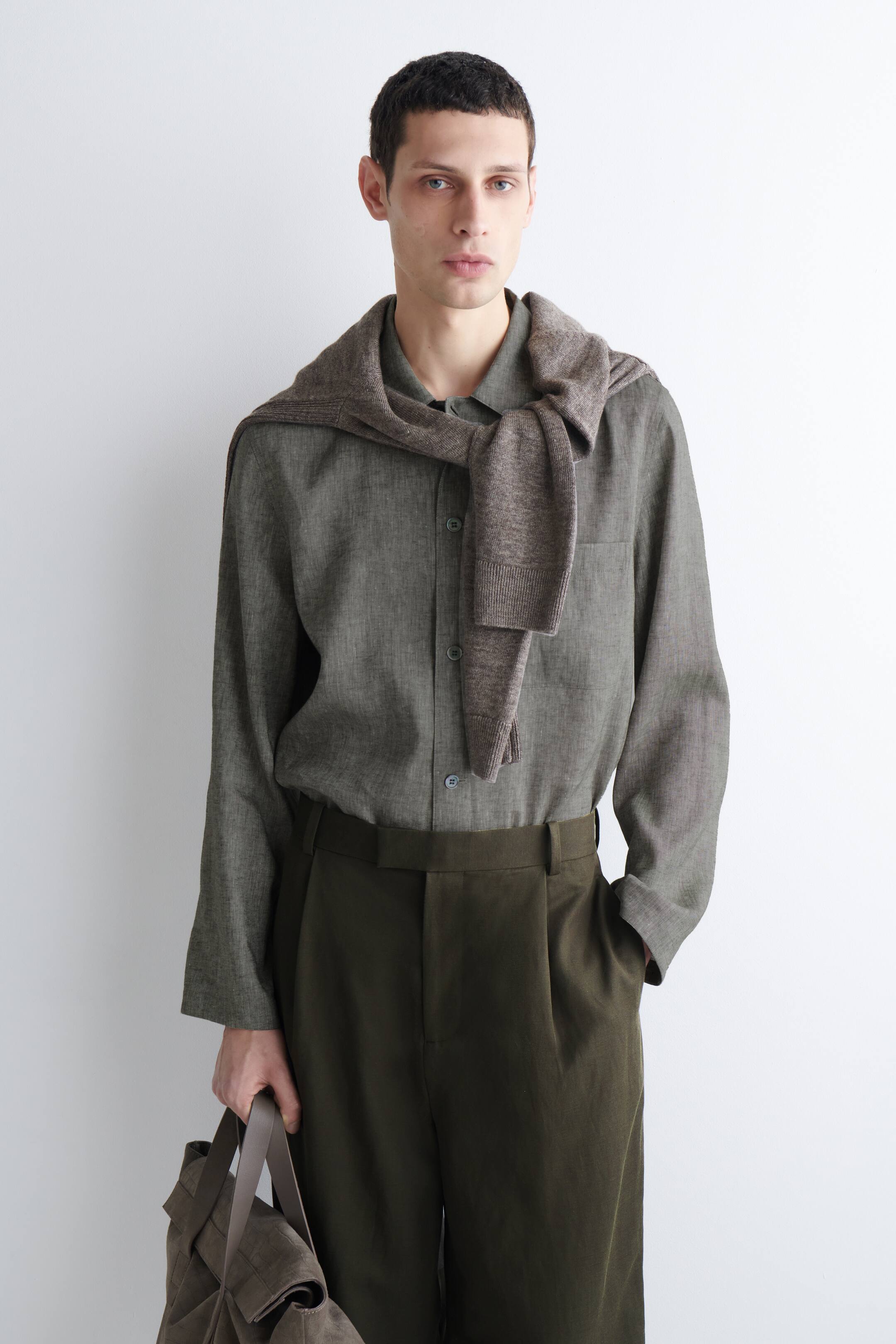 הצגת תמונה גדולה יותר: חולצה ירוקה-זית עם שרוולים ארוכים וכפתורים קדמיים נראים, עם צווארון, נלבשת עם סוודר חום מצולע המונח על הכתפיים. מכנסיים ירוקים-זית כהים עם קפלים נלבשים, בעוד תיק קניות קלוע חום כהה מוחזק.