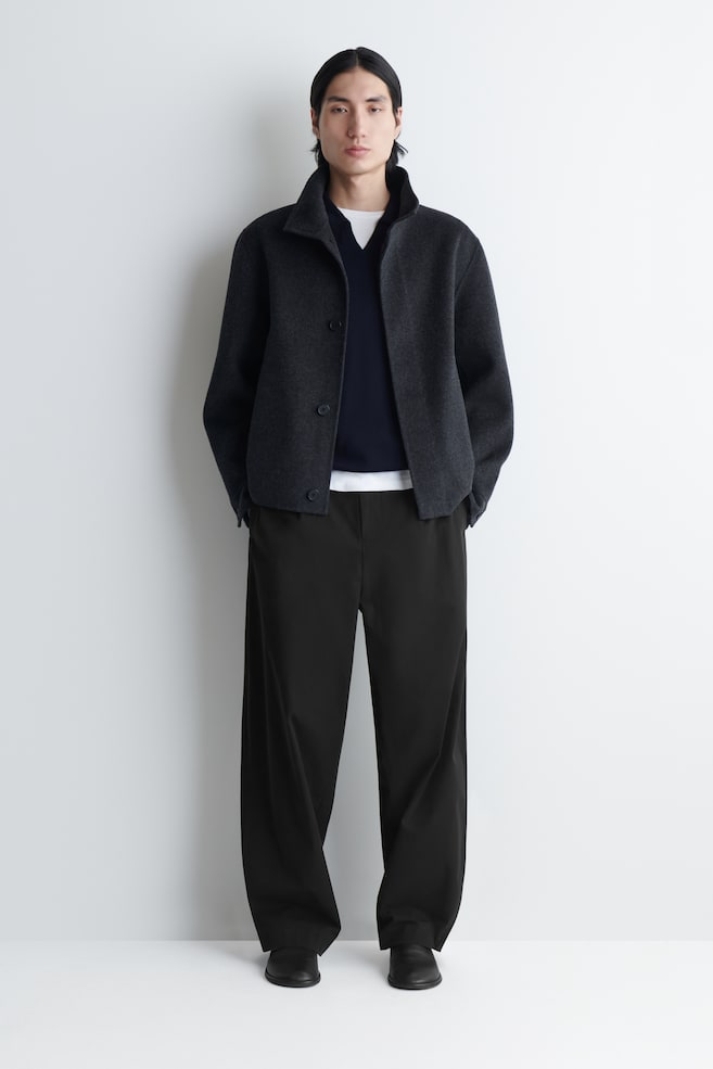 Eine dunkelgraue Jacke wird präsentiert, die mit einem Stehkragen und einem dreiknöpfigen Verschluss versehen ist. Die Seitentaschen werden genutzt, und sie ist über einem marineblauen V-Ausschnitt-Pullover und einem weißen T-Shirt geschichtet. - 1