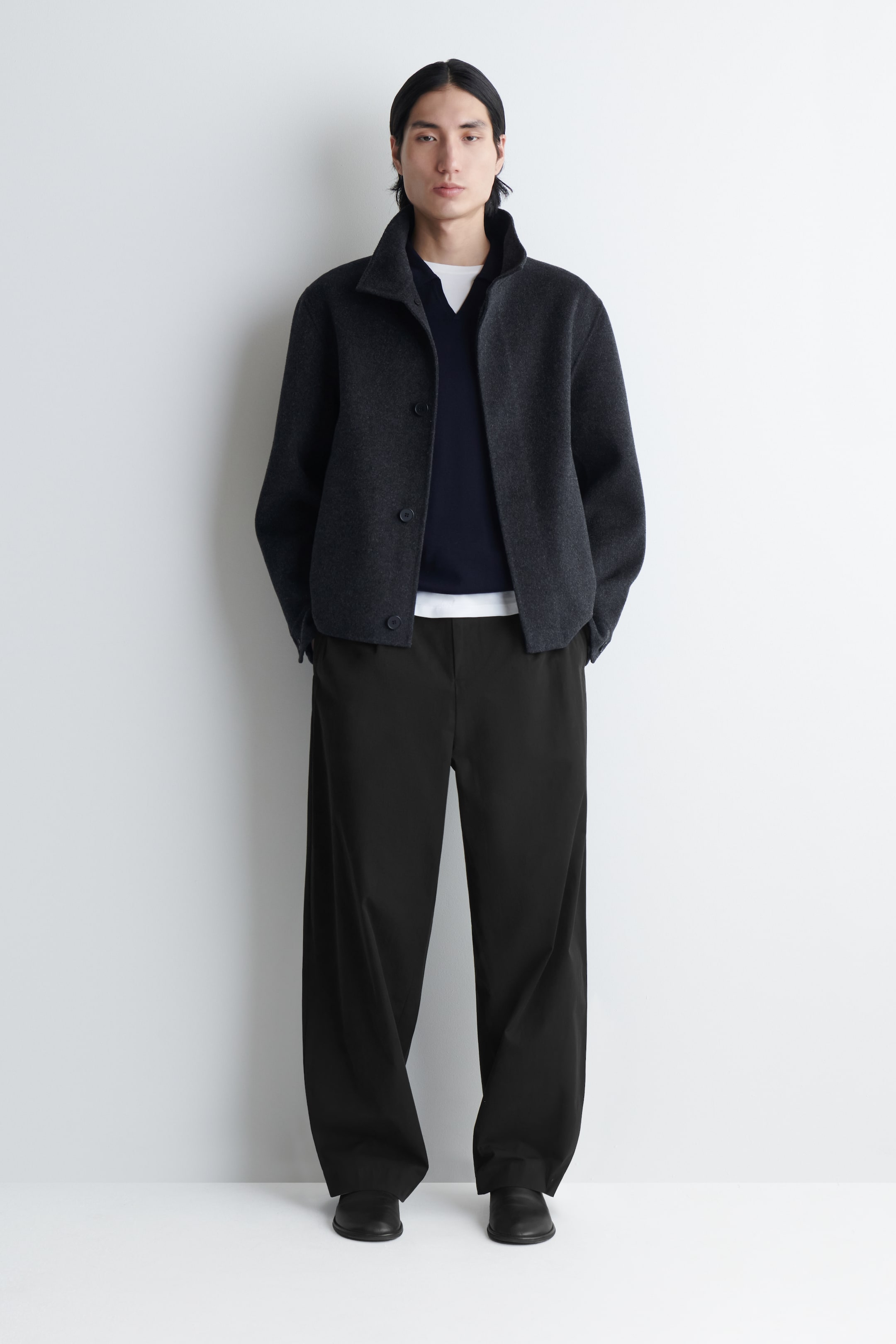 Größeres Bild ansehen: Eine dunkelgraue Jacke wird präsentiert, die mit einem Stehkragen und einem dreiknöpfigen Verschluss versehen ist. Die Seitentaschen werden genutzt, und sie ist über einem marineblauen V-Ausschnitt-Pullover und einem weißen T-Shirt geschichtet.