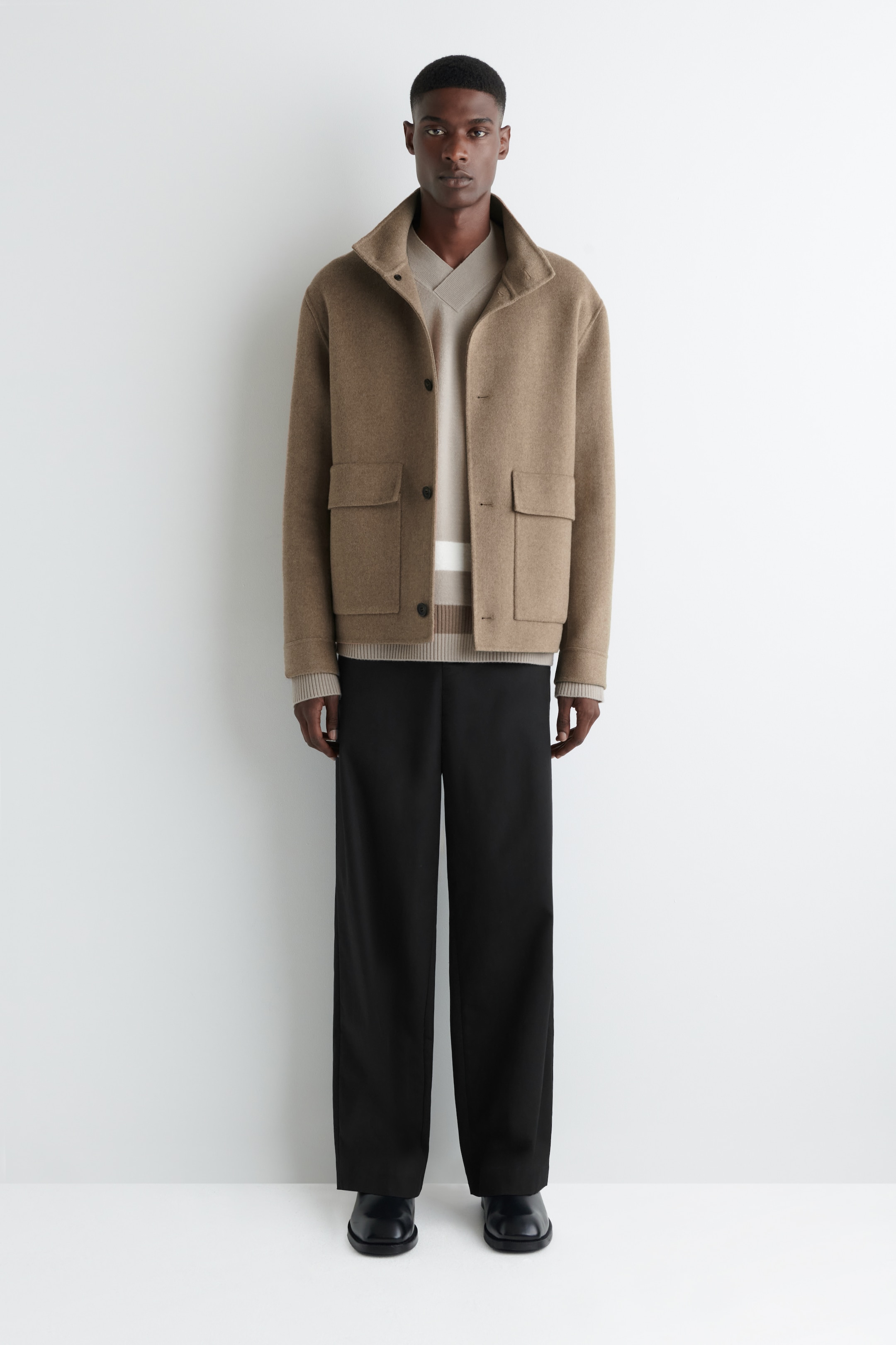 View larger image: A camel-colored, box-fit jacket is being presented from a full-front view, featuring a stand collar, button-front closure, and two flap patch pockets. It is layered over a beige V-neck sweater with a brown and cream striped hem, which is being paired with black wide-leg trousers and black square-toe shoes.