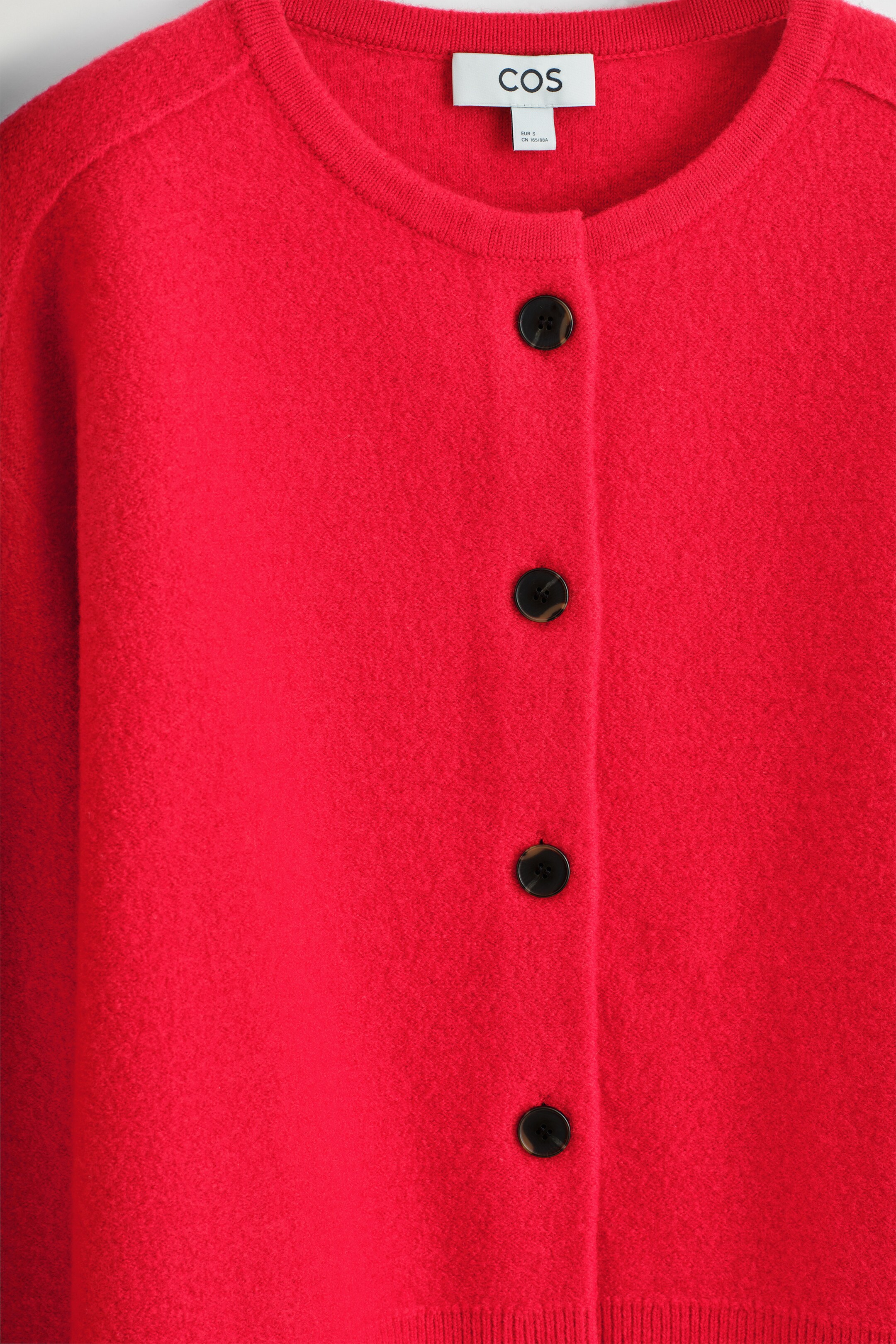Agrandir l'image: Un cardigan rouge présente son DEVANT, avec un col rond côtelé et une fermeture à boutons avec quatre boutons ronds et sombres. Le cardigan affiche une surface doucement texturée et des détails côtelés à l'ourlet, tandis que des coutures chevauchantes sont visibles aux épaules.