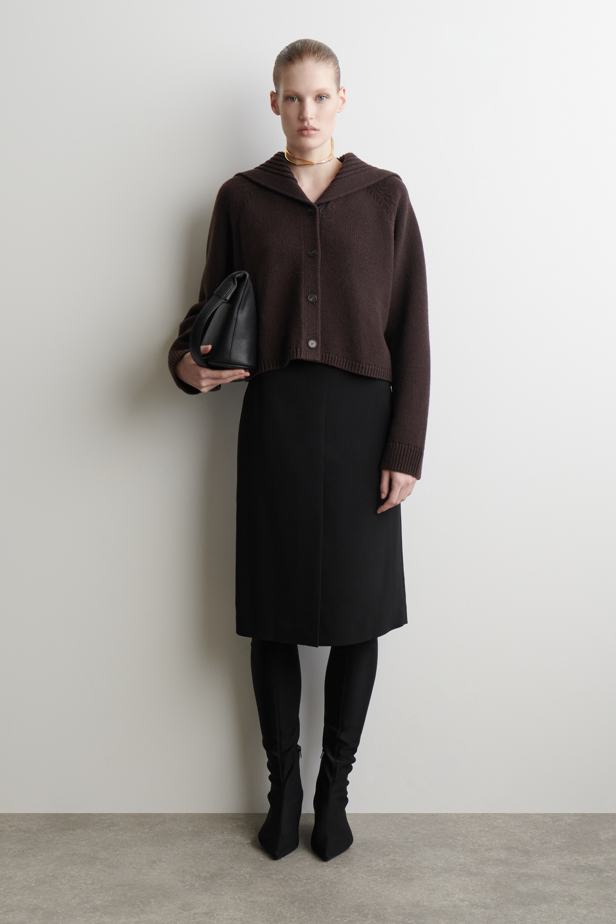 Größeres Bild ansehen: Eine dunkelbraune, kurze Strickjacke wird von vorne präsentiert, sie hat einen Schalkragen, einen Verschluss mit vier sichtbaren Knöpfen und gerippte Bündchen und Saum.