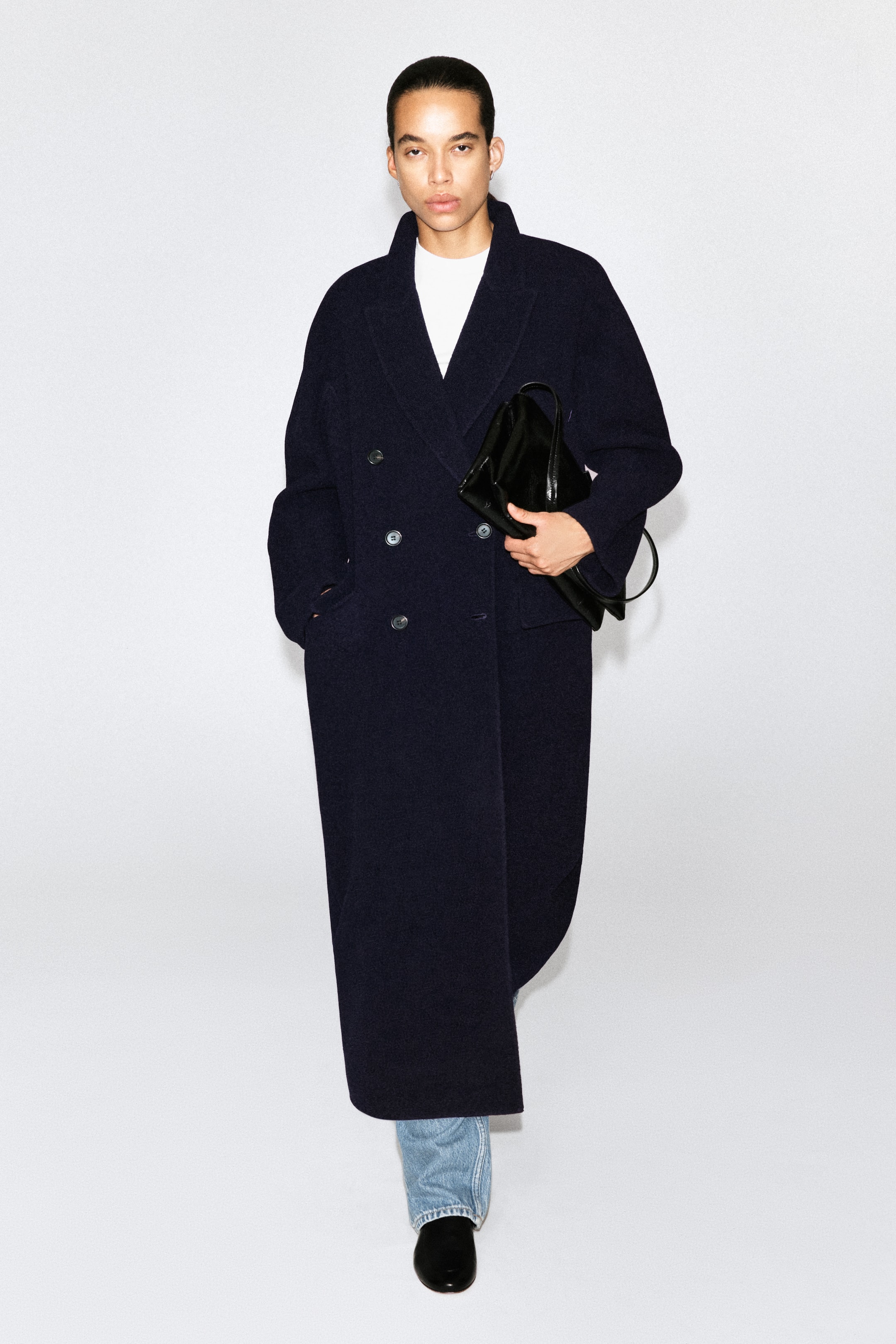 View larger image: A long, double-breasted navy wool coat is worn facing forward, showcasing its relaxed silhouette, notch lapels, and front button fastenings. The coat falls to an ankle-length hem and is styled over a white crew-neck top and light-wash jeans.