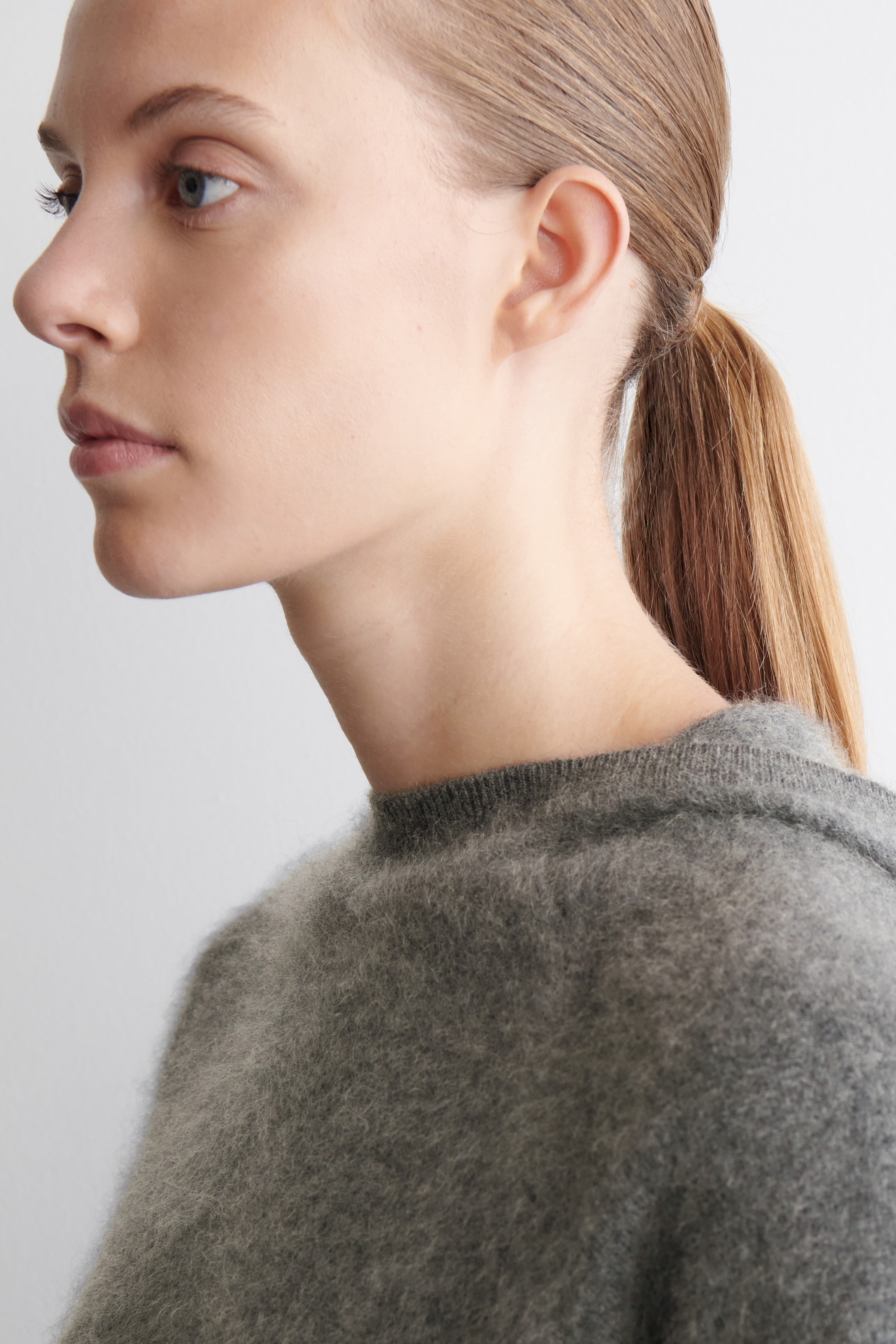 Ingrandisci l'immagine: Un maglione grigio e peloso viene presentato da un'angolazione laterale-posteriore, mettendo in risalto il suo scollo a girocollo a coste e le lunghe fibre visibili.