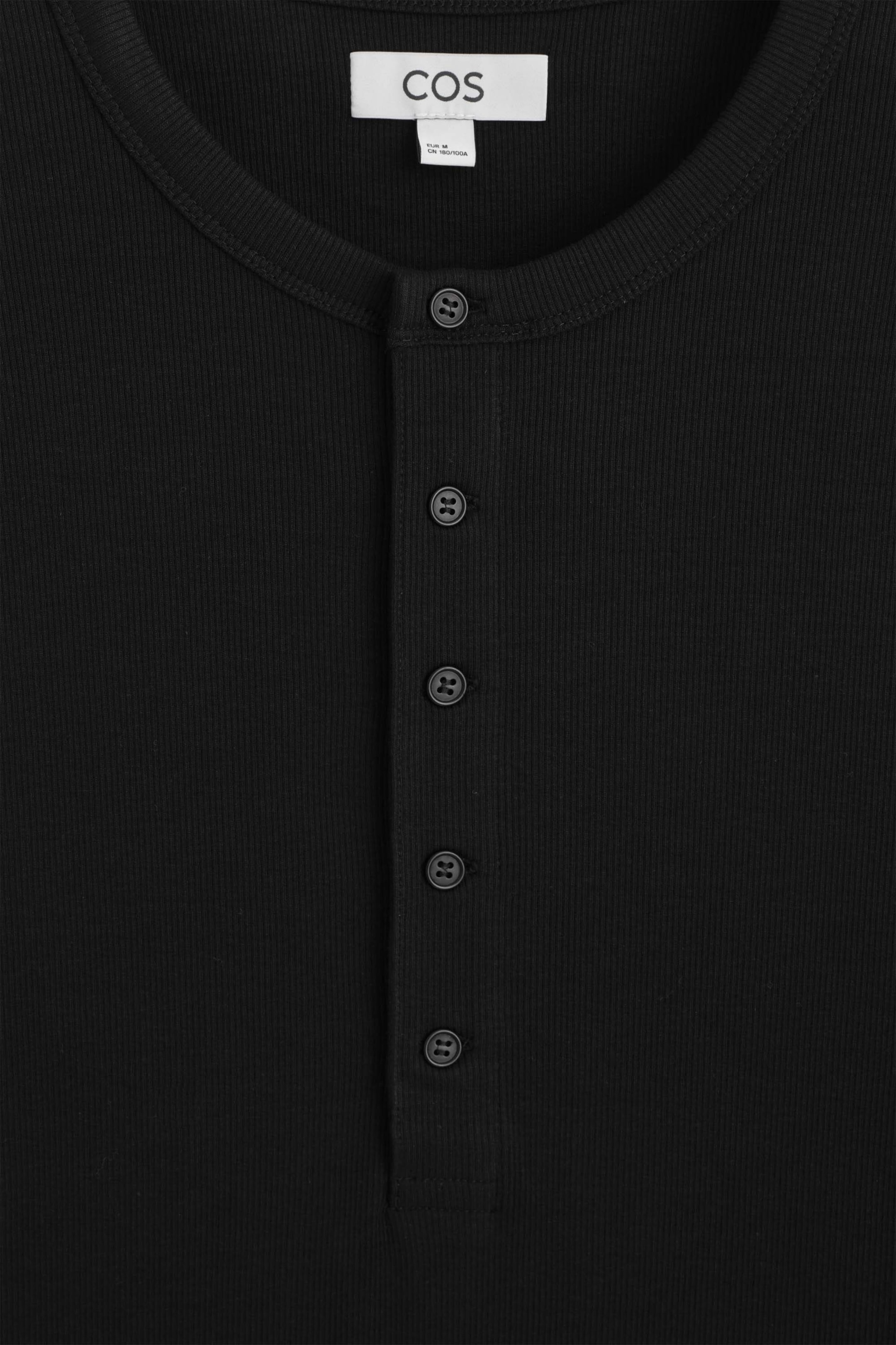The slim ribbed cotton long-sleeved henley T-shirt is shown in a close-up front view, highlighting its black ribbed-knit fabric and round neckline. A vertical placket with five black buttons runs down the centre, and a white brand label is visible on the inside collar. The slim ribbed cotton long-sleeved henley T-shirt is shown in a close-up front view, highlighting its black ribbed-knit fabric and round neckline. A vertical placket with five black buttons runs down the centre, and a white brand label is visible on the inside collar.