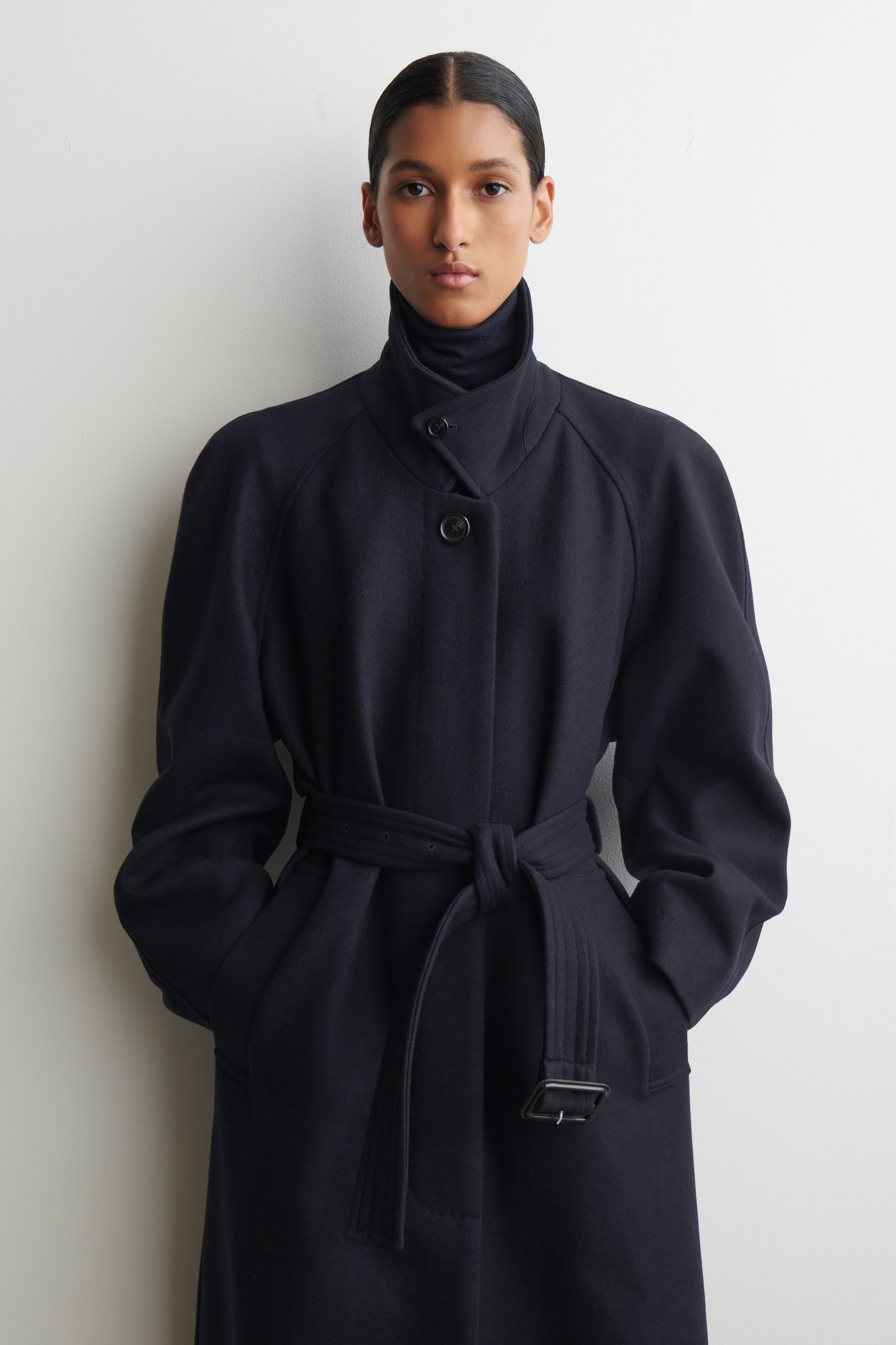 Agrandir l'image: Un manteau bleu foncé est présenté de face, avec un col montant fermé par des boutons sur un col roulé foncé, avec deux boutons visibles sur sa patte de boutonnage avant. Il présente des manches raglan volumineuses et une ceinture en tissu assorti nouée à la taille, avec les mains reposant dans ses poches latérales.