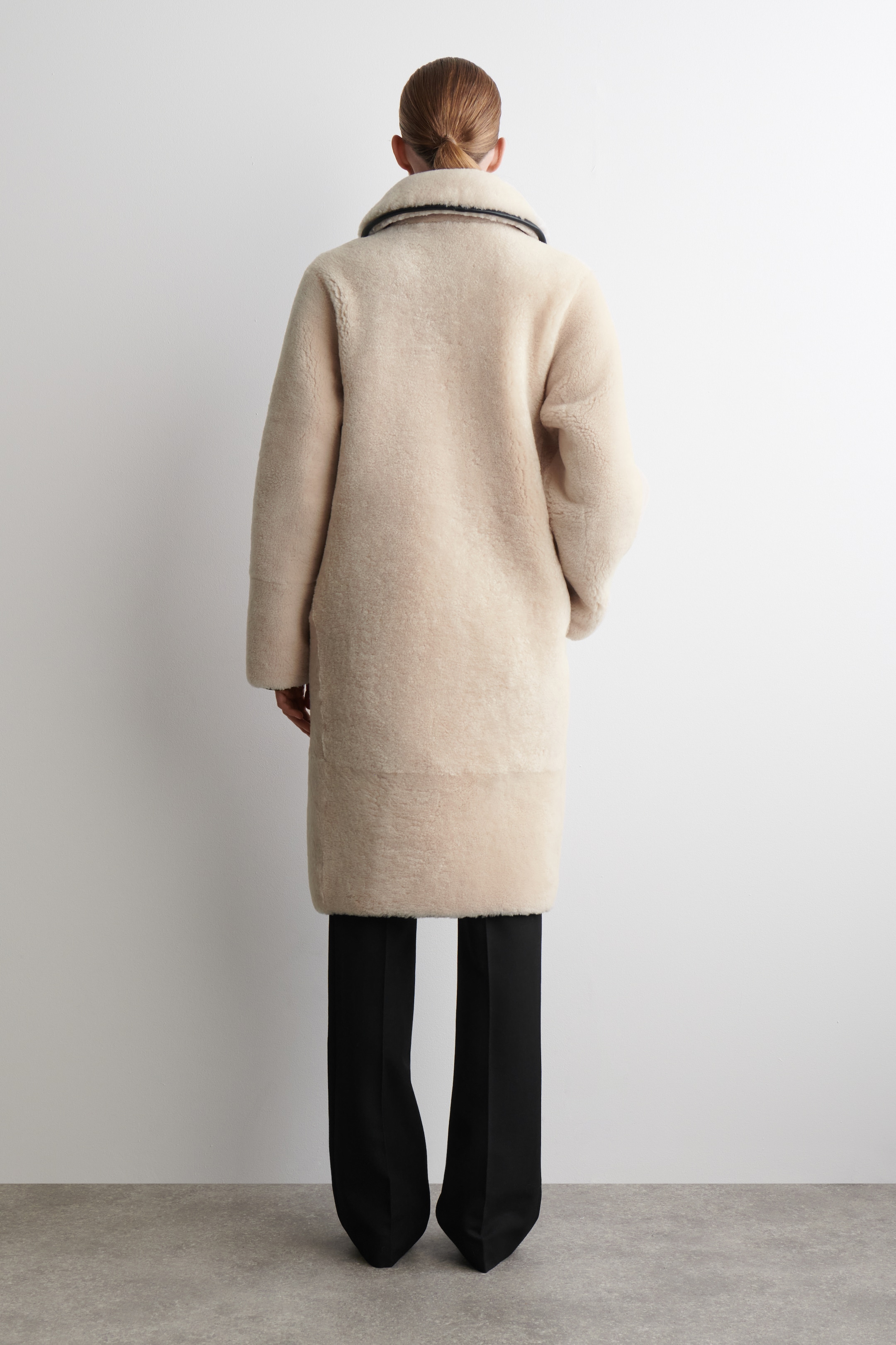 Agrandir l'image: Un manteau en shearling de couleur claire est présenté de dos. Il présente un large col plié avec une bordure sombre et des manches longues et amples, et s'étend sur un pantalon noir à jambes larges.