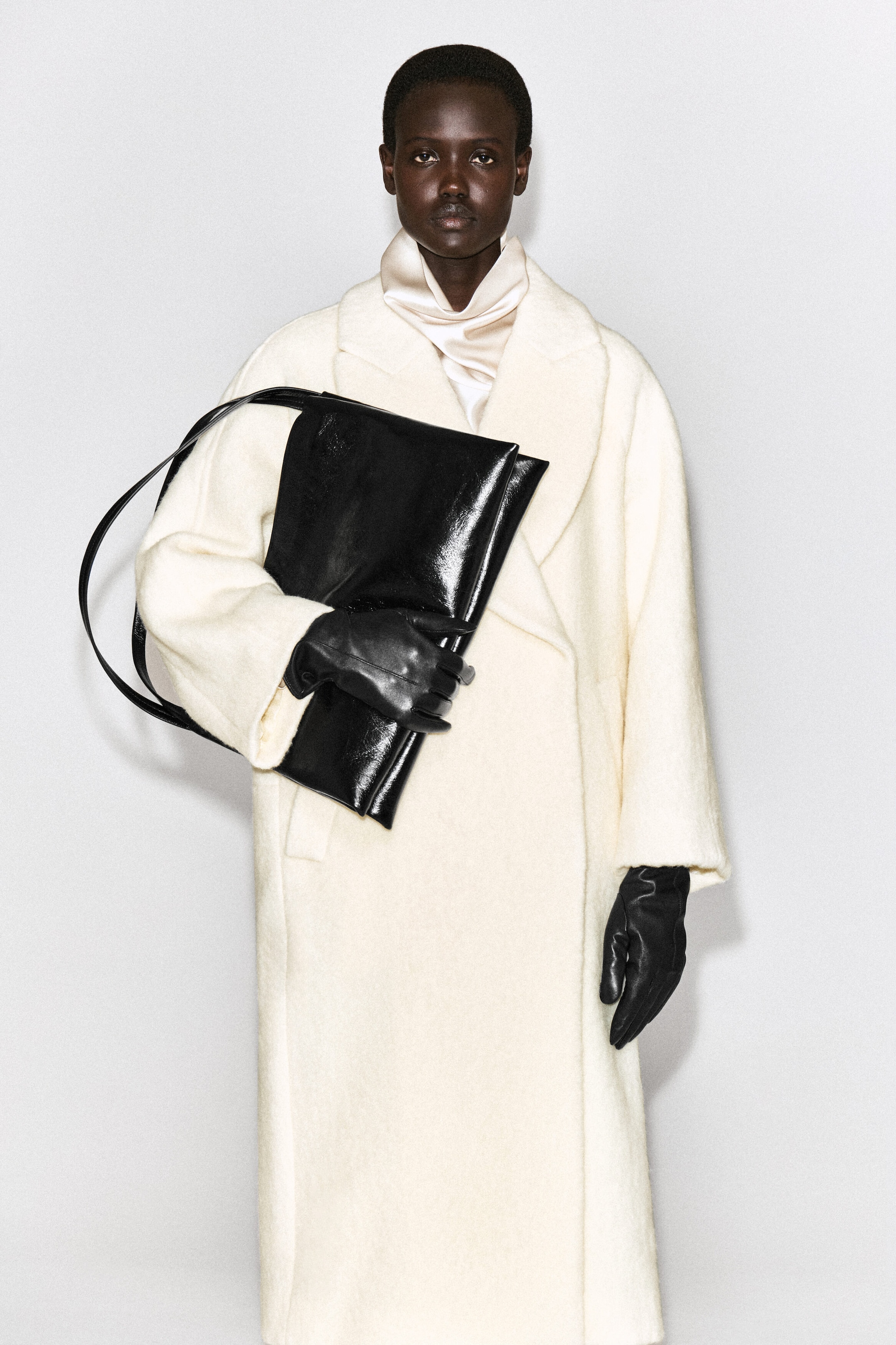 Un grand sac en cuir noir brillant est porté sous le bras par le mannequin, qui le tient d’une main gantée de noir. Le sac est doté d’une bandoulière fine et est agencé avec un long manteau texturé blanc cassé. Un grand sac en cuir noir brillant est porté sous le bras par le mannequin, qui le tient d’une main gantée de noir. Le sac est doté d’une bandoulière fine et est agencé avec un long manteau texturé blanc cassé.