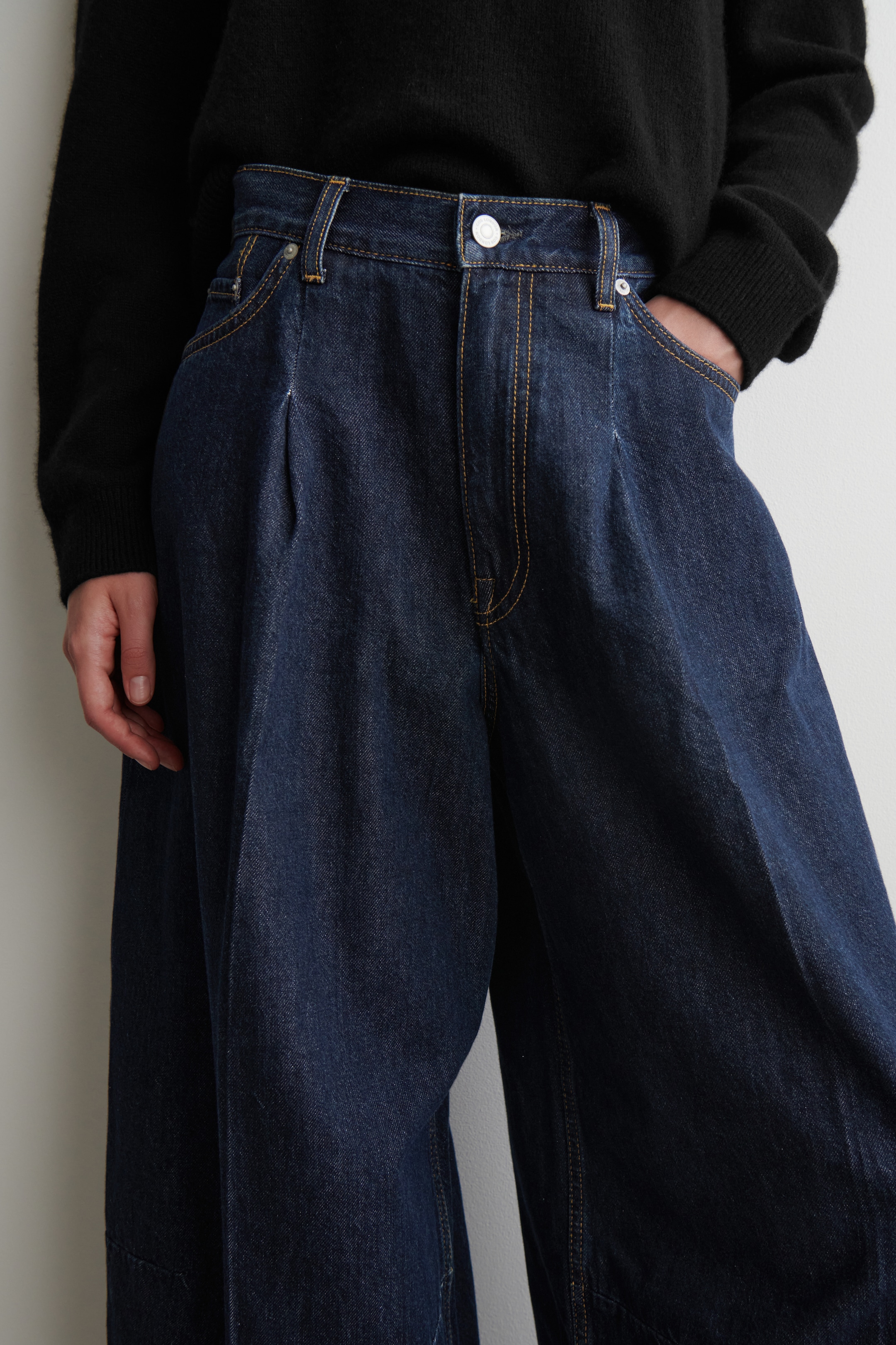 Visualizza immagine più grande: I pantaloni a gamba larga in denim indaco scuro presentano cuciture a contrasto gialle, una singola piega su ciascuna gamba e tasche laterali, mostrando una vestibilità rilassata e ampia. Una mano è appoggiata in una tasca, evidenziandone il design.