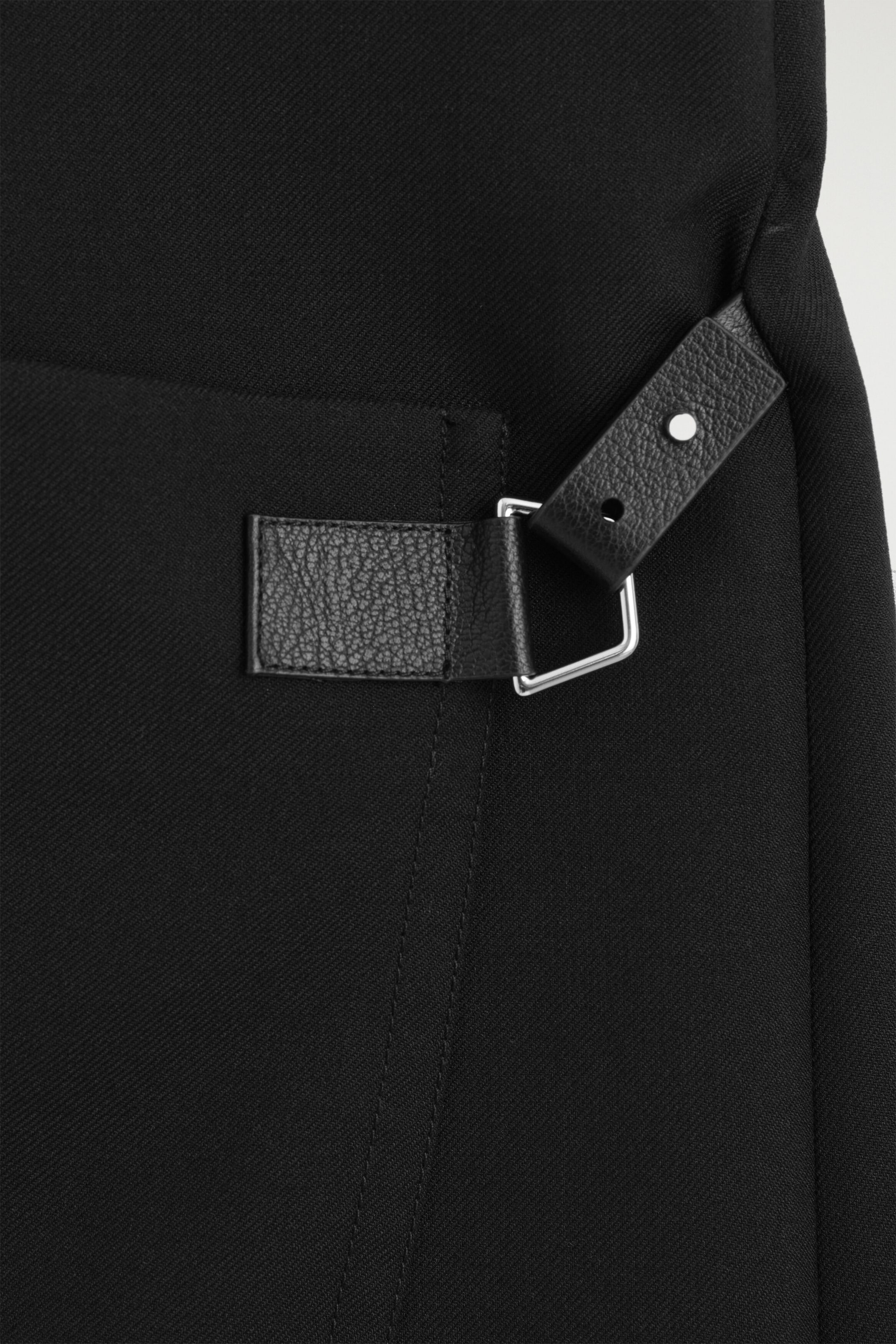 The belt-detail double-faced wool midi dress is shown in a close-up of the side waist detail, featuring a textured black leather strap looped through a silver-tone metal ring. The strap showcases a silver-tone metal stud and adjustment holes against the diagonally woven black wool fabric.