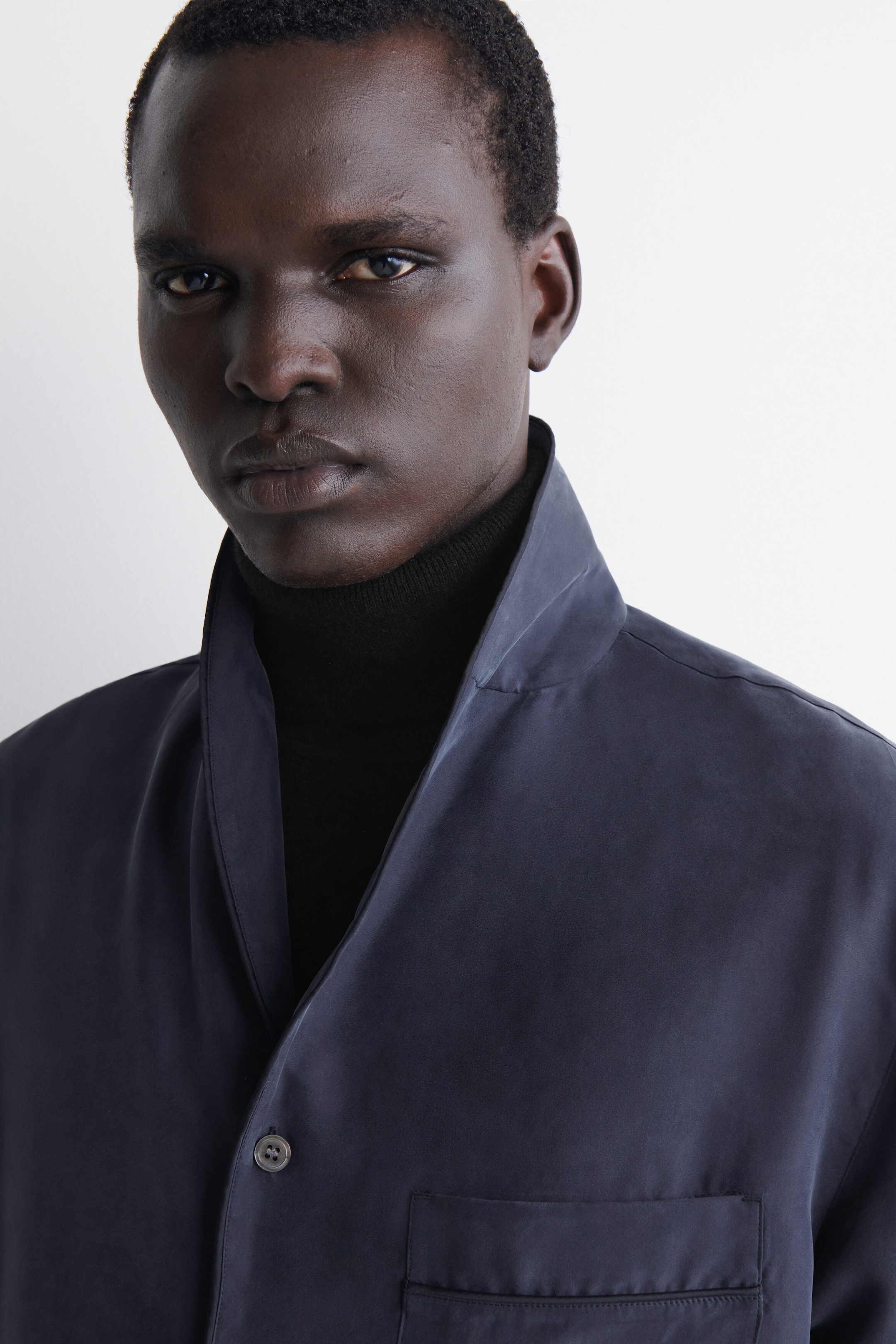 A dark grey silk shirt is worn over a black roll-neck jumper, showcasing its soft shawl collar and fluid drape. The front close-up view shows a chest pocket with piped trim and a single button fastening. A dark grey silk shirt is worn over a black roll-neck jumper, showcasing its soft shawl collar and fluid drape. The front close-up view shows a chest pocket with piped trim and a single button fastening.
