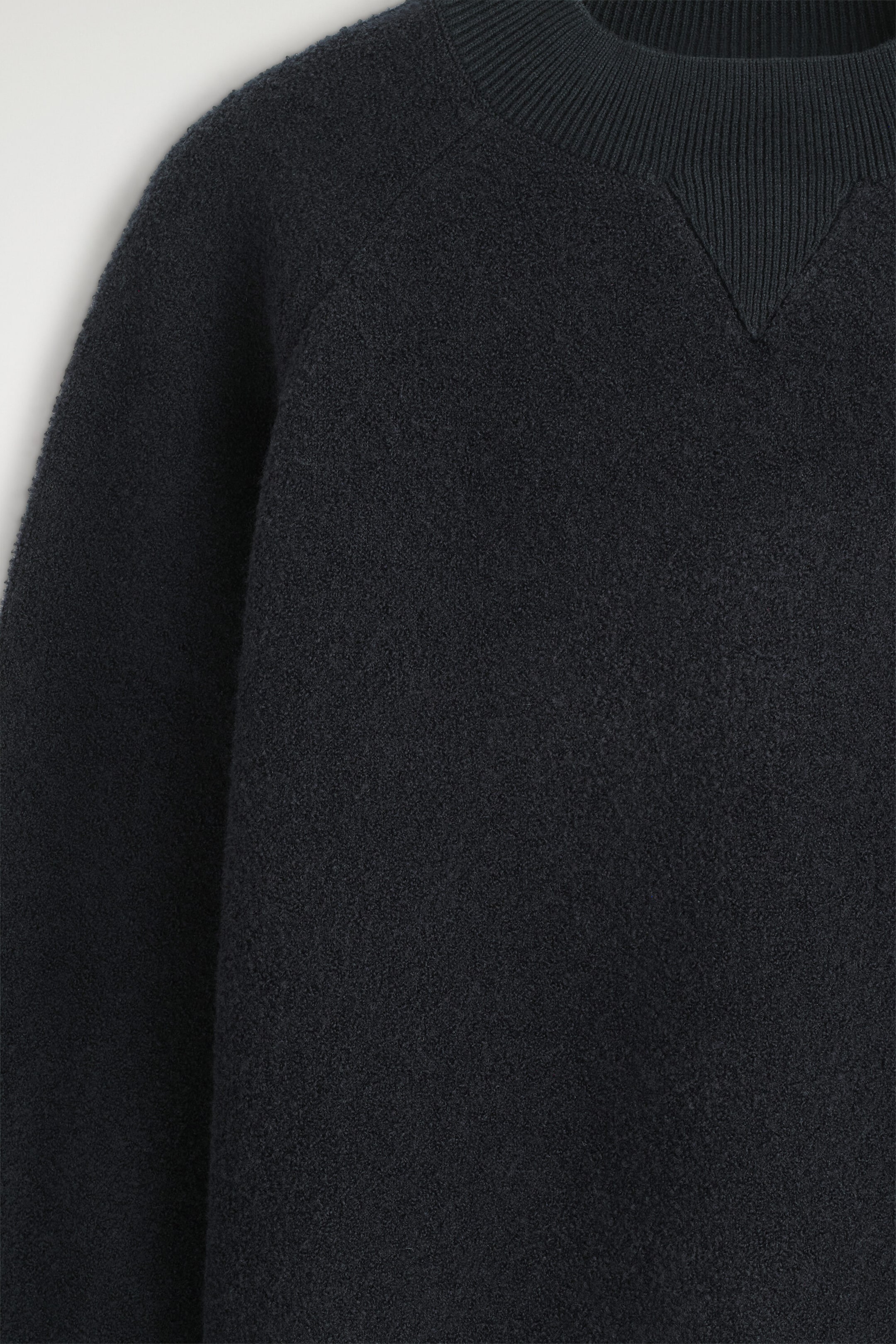 A dark navy bouclé jumper is shown in close-up, highlighting its textured knit fabric, with a ribbed mock neck, tonal V-insert, and raglan sleeve seam. A dark navy bouclé jumper is shown in close-up, highlighting its textured knit fabric, with a ribbed mock neck, tonal V-insert, and raglan sleeve seam.