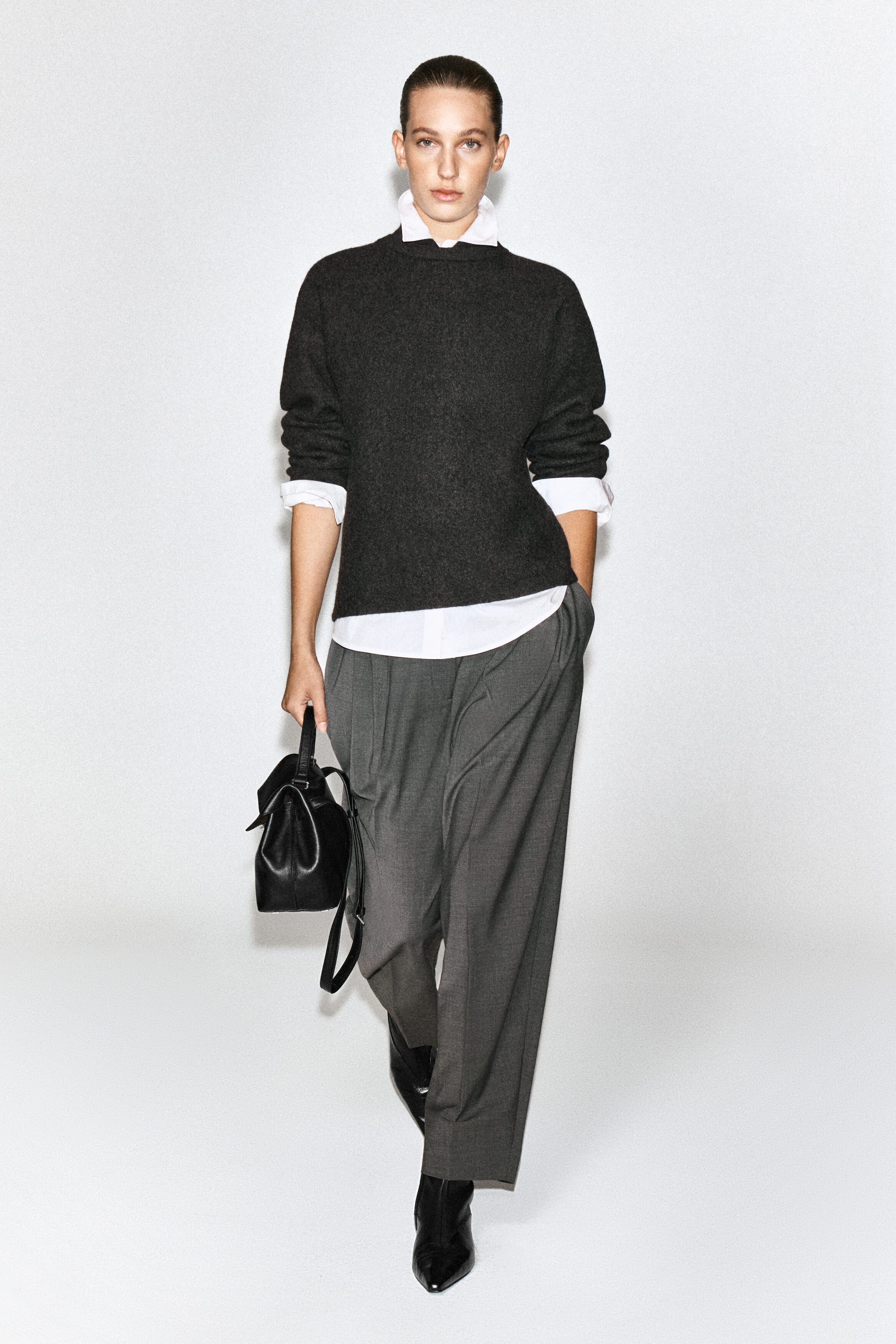 The model is wearing a dark gray boiled wool sweater over a white collared shirt, with the white collar, cuffs, and hem visibly layered underneath. The outfit is styled with wide-leg gray trousers, and the model is carrying a black leather handbag.