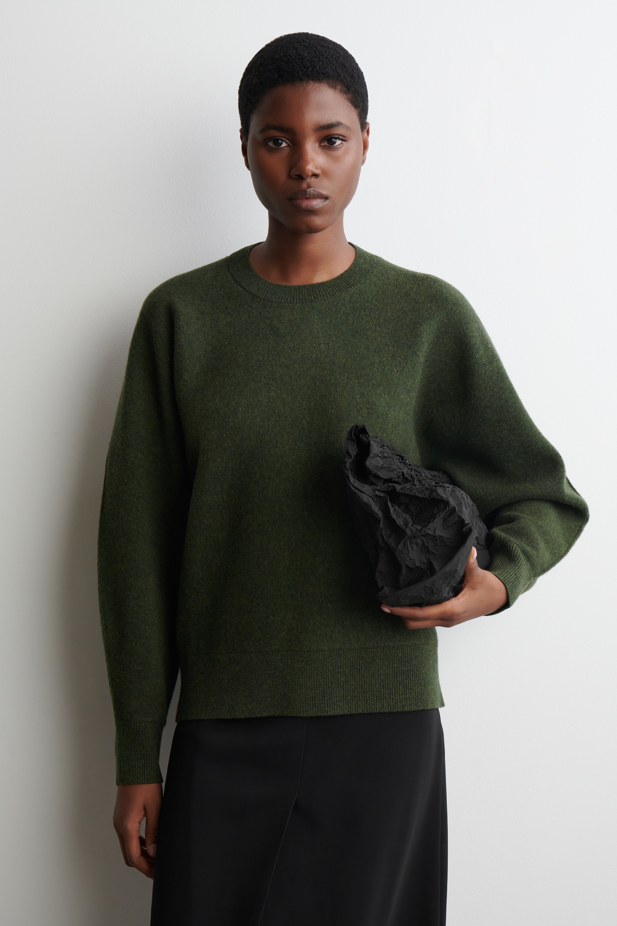 Grösseres Bild ansehen: Ein dunkelgrüner Pullover mit Rundhalsausschnitt wird von vorne gezeigt, mit einer lockeren Passform und überschnittenen Schultern. Er hat einen gerippten Rundhalsausschnitt, gerippte Bündchen und einen gerippten Saum, die alle ein weiches, strukturiertes Strickmuster aufweisen.