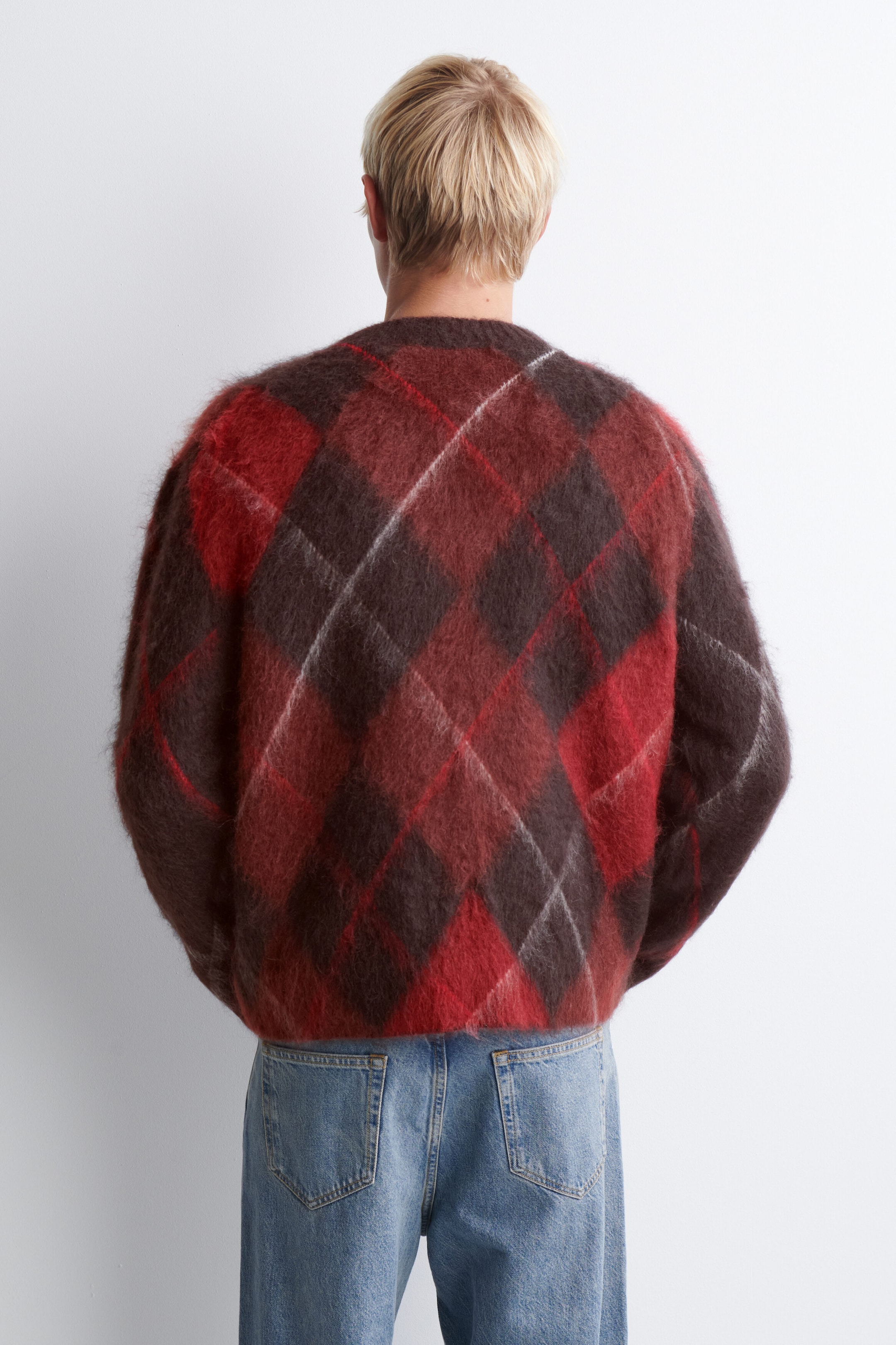 Dieser flauschige Strickpullover mit einem grossen Argyle-Muster in Rot, Dunkelbraun und Weiss ist von hinten gezeigt, um seine lockere Passform und die weiche, aufgeraute Struktur hervorzuheben. Der Pullover sitzt locker auf der Taille über hellblauen Jeans in Waschung.