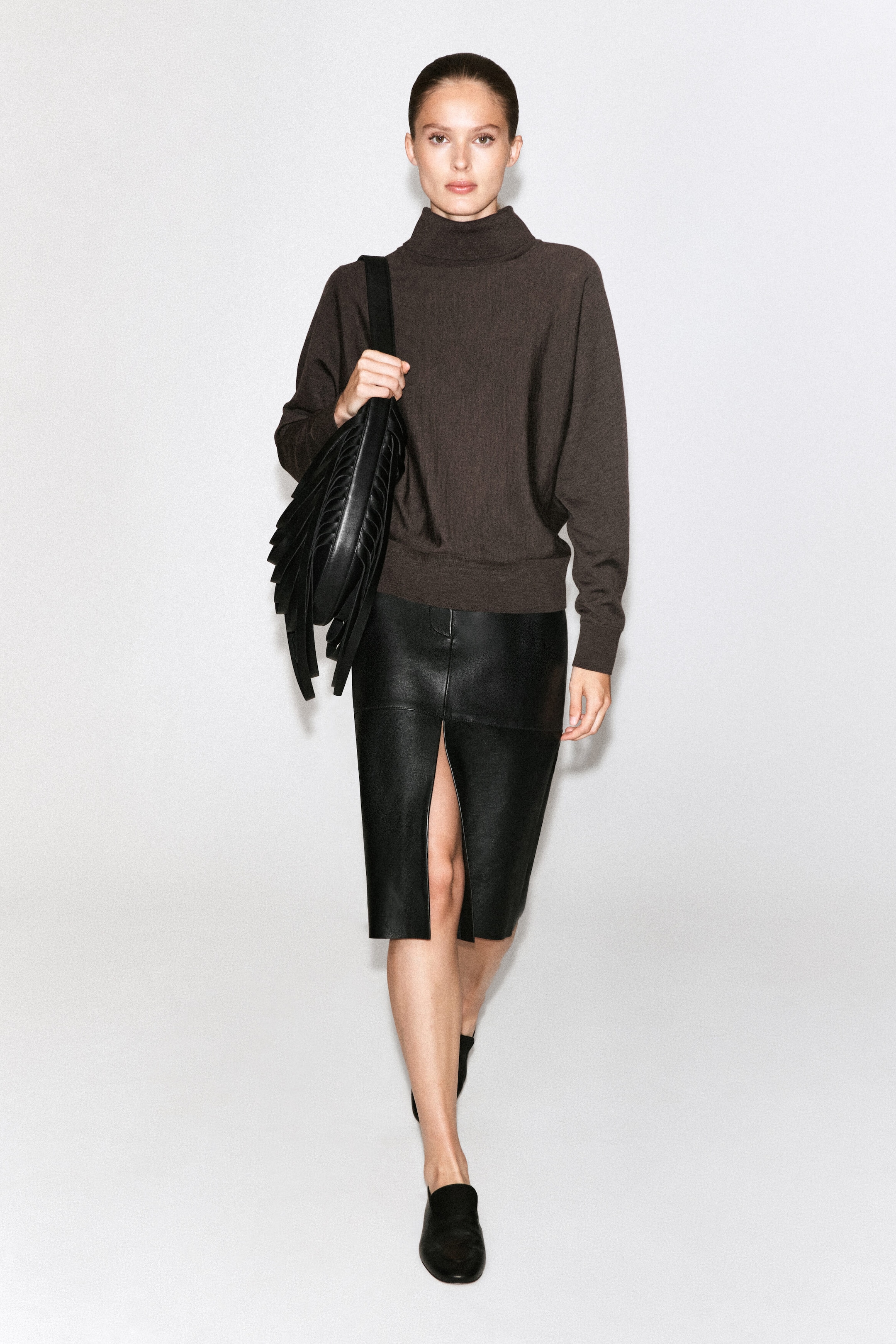 A dark brown, long-sleeved roll-neck jumper is being worn by the model, paired with a high-waisted black leather skirt featuring a front slit. A black leather shoulder bag with long fringe detailing is being carried on one shoulder, completing the ensemble. A dark brown, long-sleeved roll-neck jumper is being worn by the model, paired with a high-waisted black leather skirt featuring a front slit. A black leather shoulder bag with long fringe detailing is being carried on one shoulder, completing the ensemble.