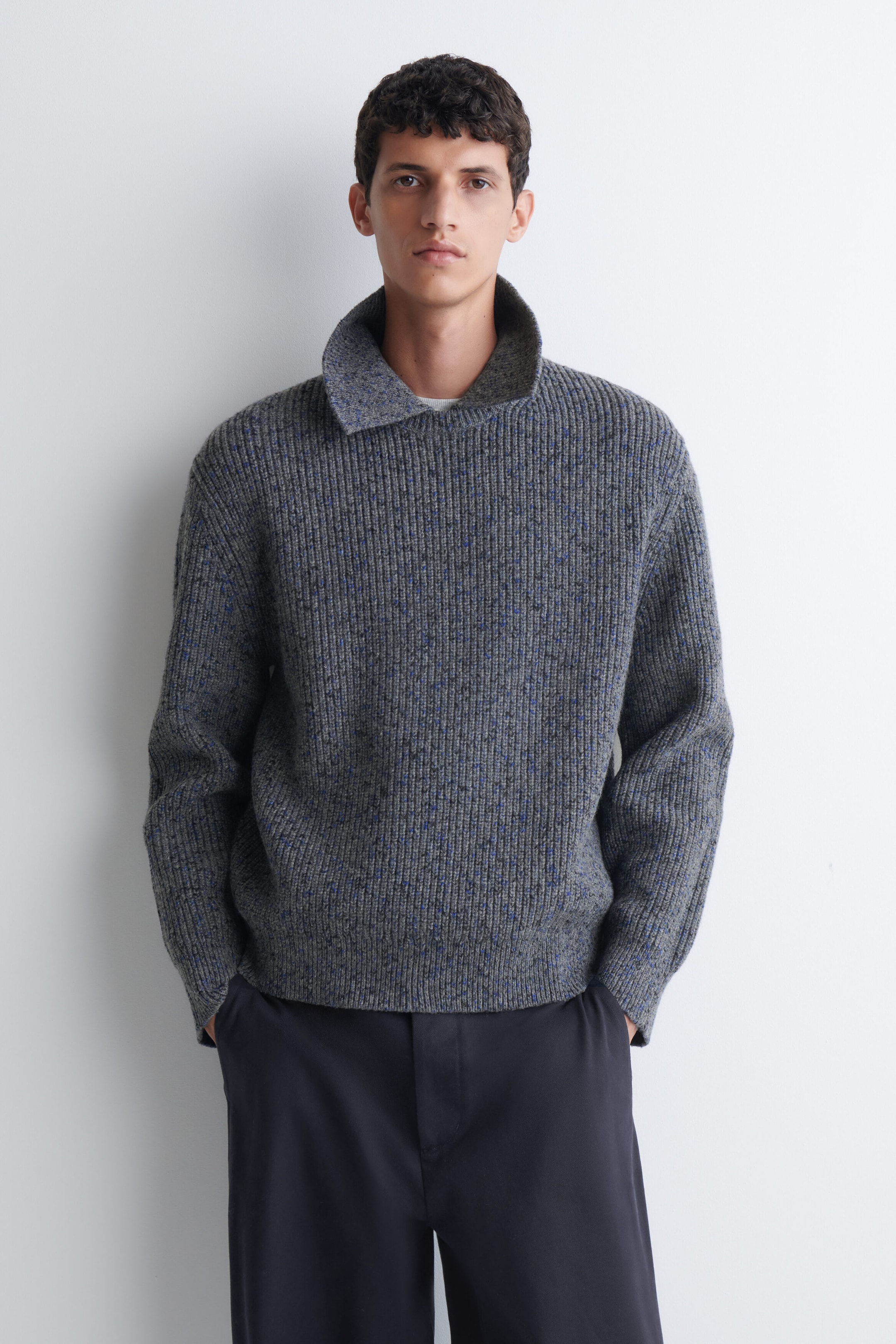 A grey ribbed-knit sweater with blue speckled details is being worn by the model. The sweater features a high, open collar and is styled with dark trousers. A grey ribbed-knit sweater with blue speckled details is being worn by the model. The sweater features a high, open collar and is styled with dark trousers.