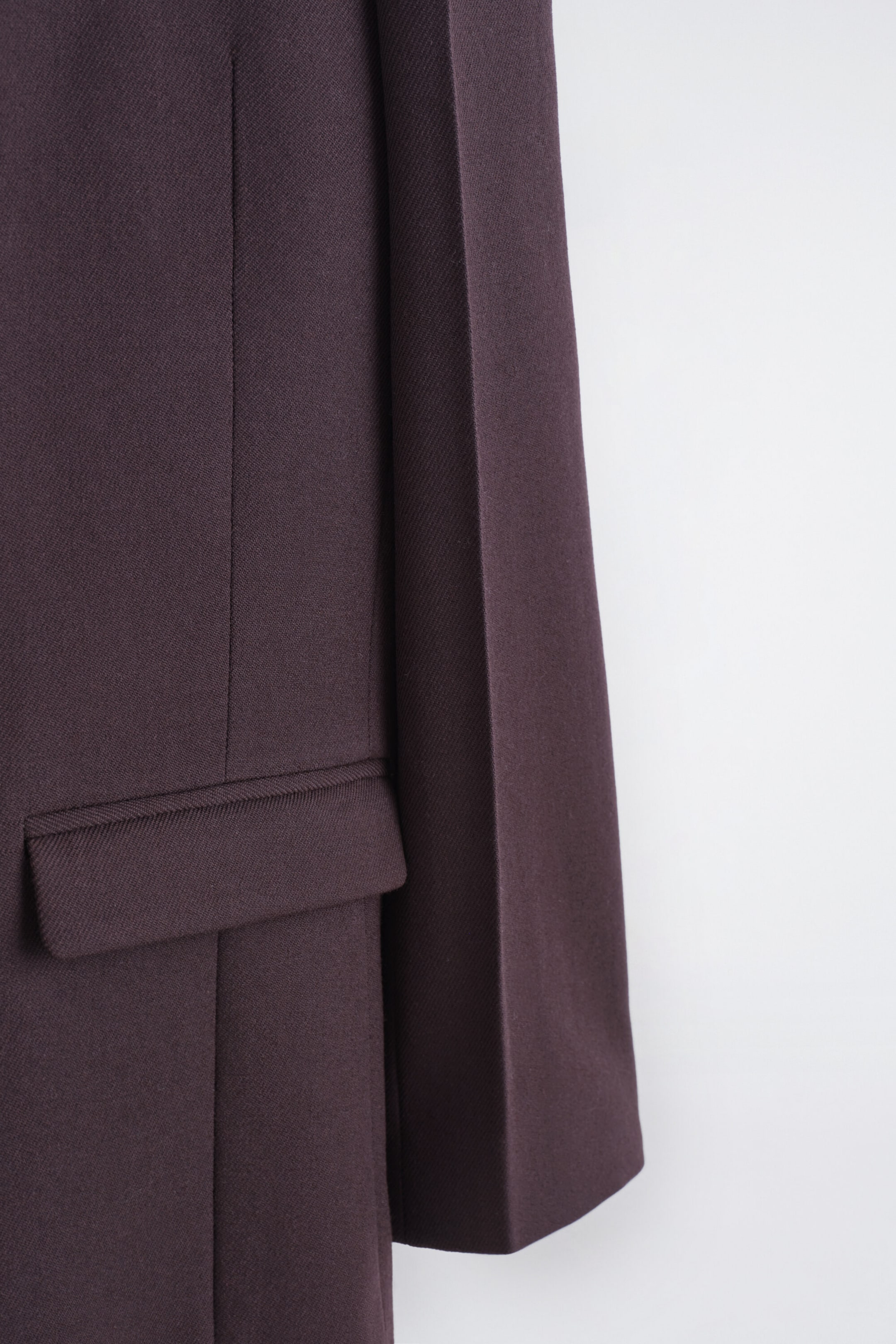 A dark purple jacket is shown from the side, revealing a horizontal flap pocket and a vertical seam above it. The fabric shows a subtle diagonal texture, and a straight sleeve extends to the right. A dark purple jacket is shown from the side, revealing a horizontal flap pocket and a vertical seam above it. The fabric shows a subtle diagonal texture, and a straight sleeve extends to the right.