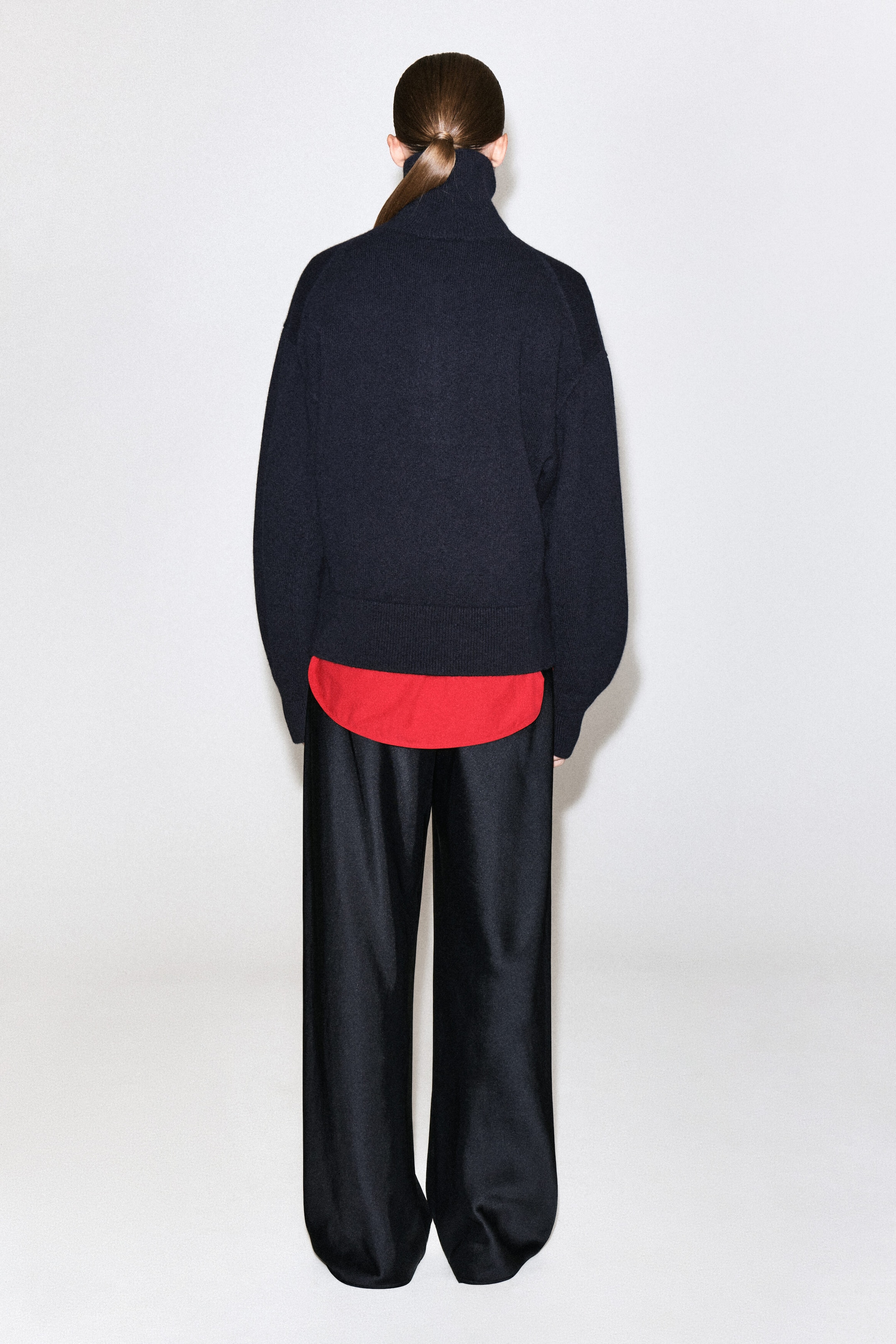 A dark navy knitted jumper with a high collar is being worn by the model, shown from the back. The jumper features raglan sleeves and a ribbed hem, and is styled in layers over a red shirt and paired with black wide-leg trousers.
