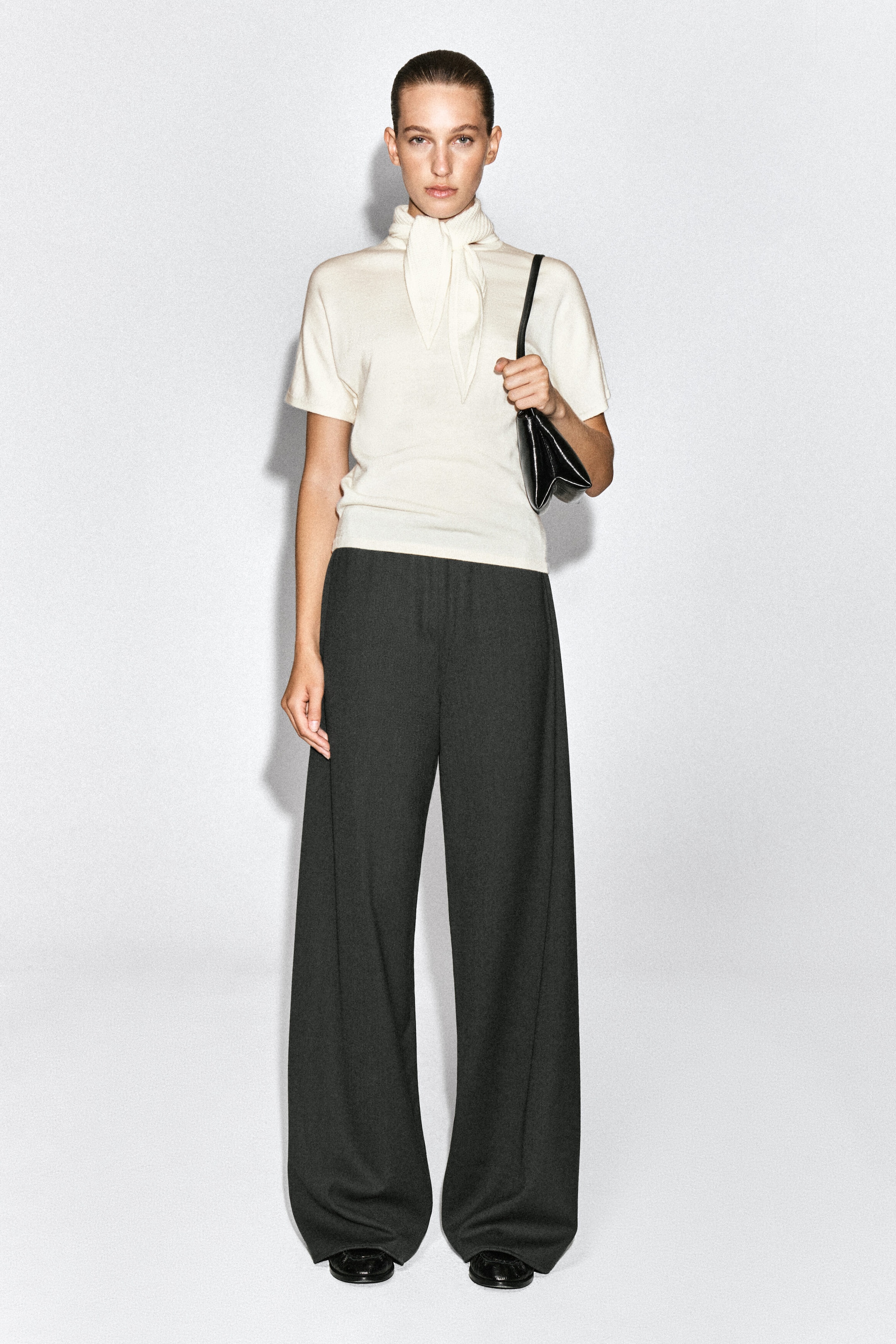 The model is wearing an off-white merino wool-blend T-shirt with short sleeves and an integrated neck scarf tied at the front. The top is styled with dark wide-leg trousers, and the model is carrying a small black shoulder bag.
