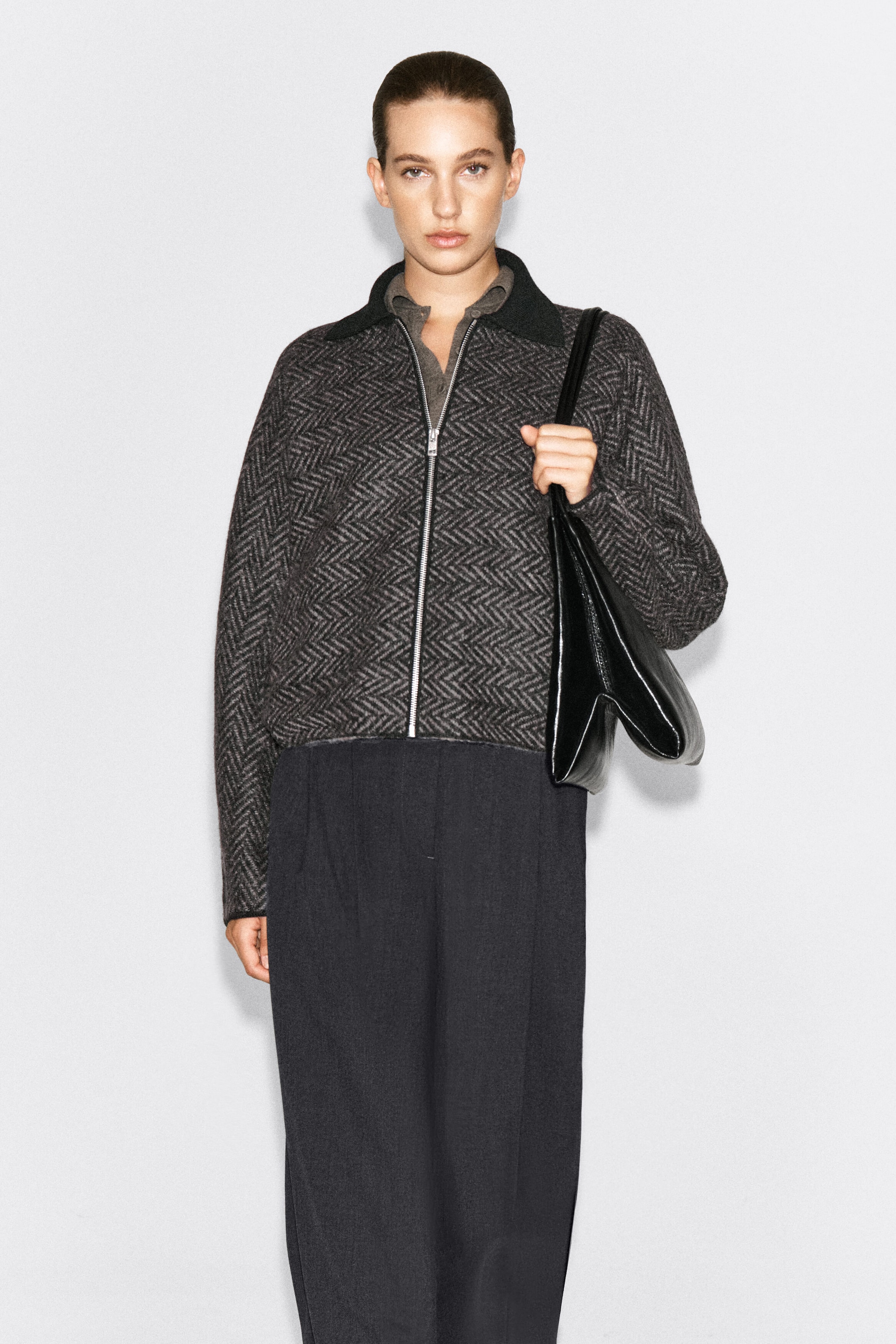 Le mannequin porte une veste en tricot à chevrons gris et noir, dotée d’un col côtelé foncé et d’une fermeture éclair frontale argentée. La veste est agencée avec un pantalon foncé et un sac à bandoulière en cuir noir.