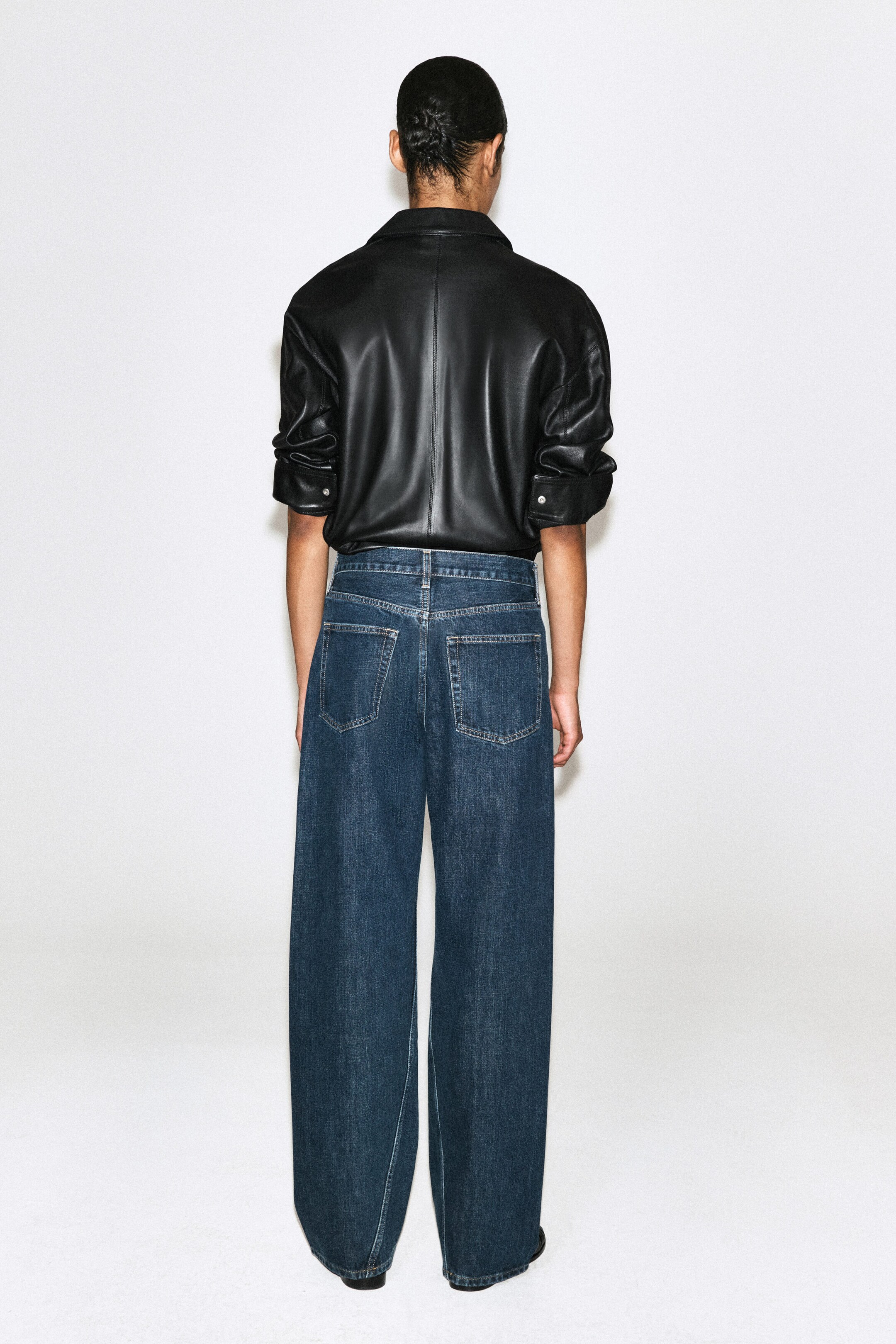 These dark blue wide-leg denim jeans, shown from the back, feature a relaxed fit with classic rear patch pockets. They are styled with a tucked-in black leather shirt featuring short sleeves and buttoned cuffs. These dark blue wide-leg denim jeans, shown from the back, feature a relaxed fit with classic rear patch pockets. They are styled with a tucked-in black leather shirt featuring short sleeves and buttoned cuffs.
